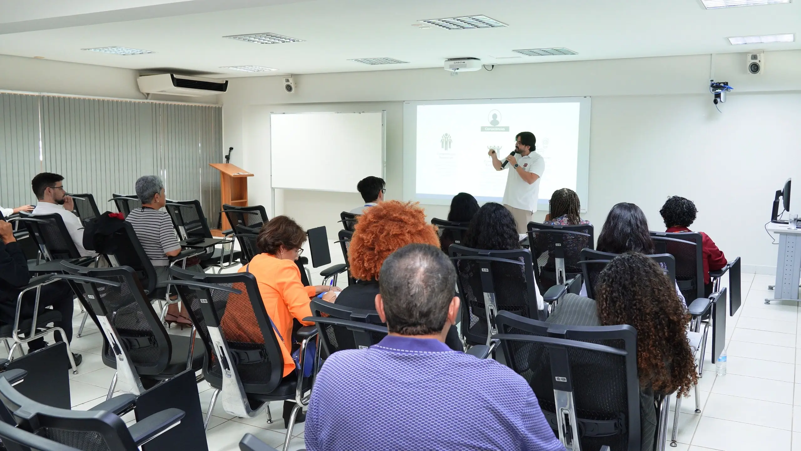 Vista da parte traseira de uma plateia em uma sala de treinamento. À frente, o palestrante e professor Thiago Dias Costa faz uma apresentação ao lado de uma tela de projeção com gráficos. 