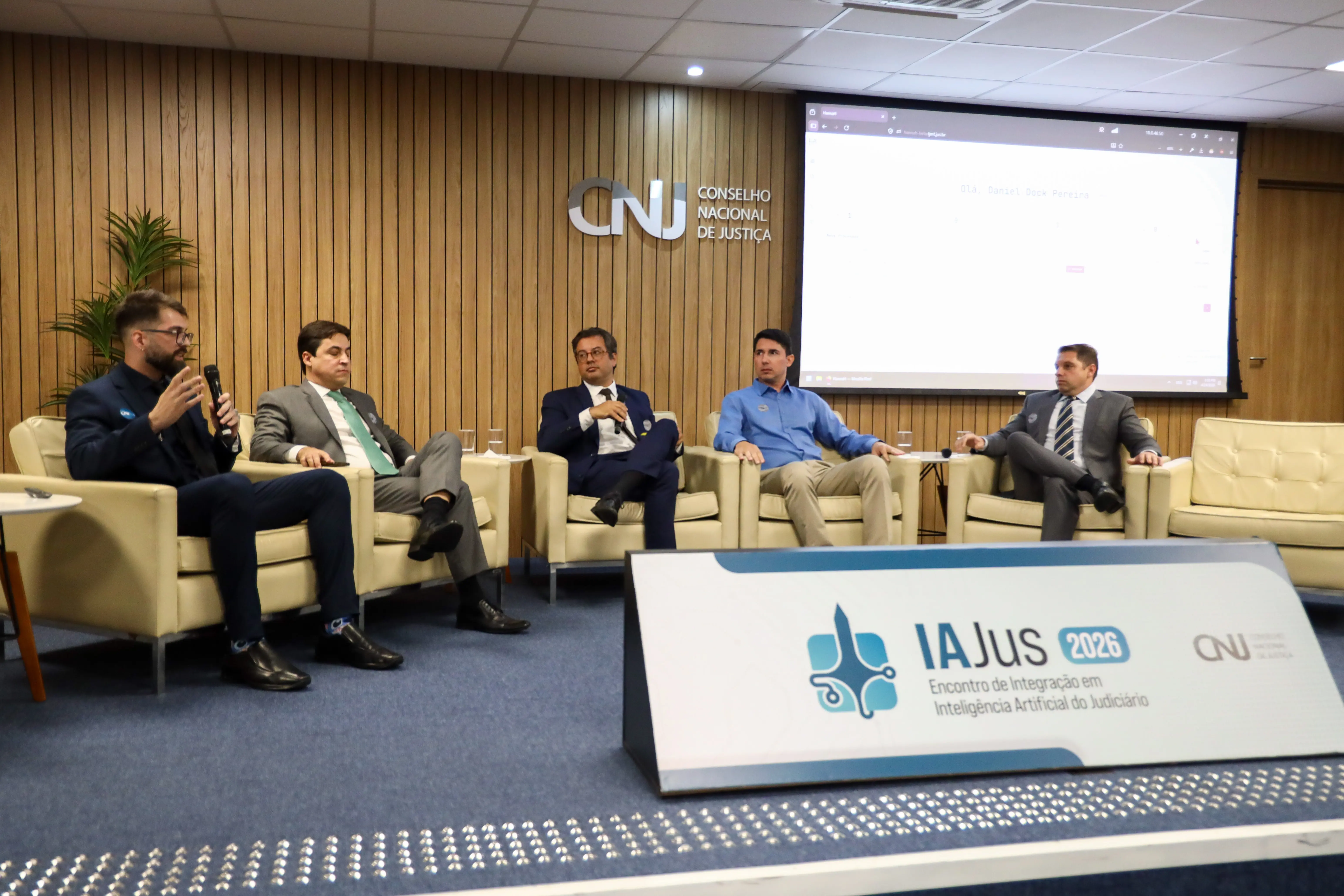 Cinco homens participam de um painel no evento IAJus 2026, no CNJ. Sentados em poltronas beges diante de uma tela de projeção, eles discutem Inteligência Artificial no Judiciário.