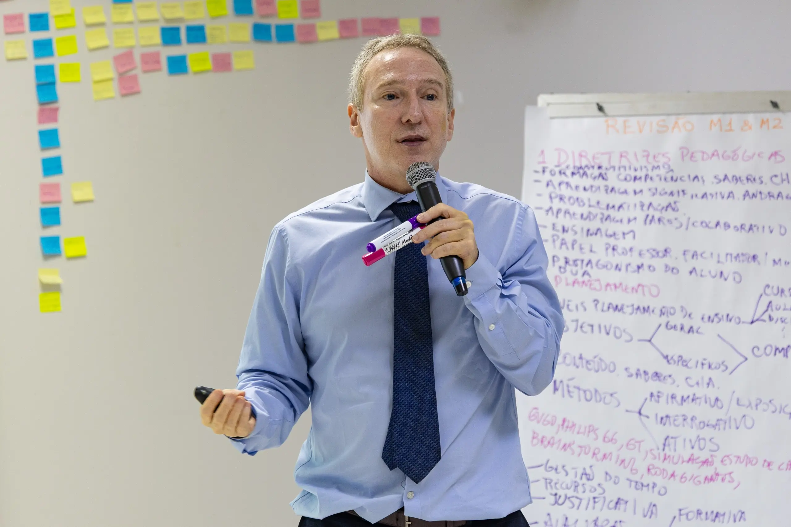 Homem branco sorridente de cabelos curtos escuros, vestindo terno cinza, camisa branca e gravata vermelha, posa à frente de uma parede branca. Ao fundo, diversos post-its coloridos estão organizados em colunas e linhas, indicando um painel de planejamento ou lousa de atividades.