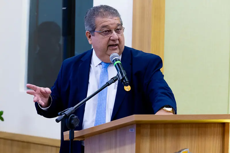Foto horizontal colorida, em plano médio, que mostra o procurador de justiça Paulo Prado falando ao microfone, no púlpito de um auditório. Ele é um senhor branco, obeso, de cabelos grisalhos, usando camisa branca, terno azul marinho, gravata azul clara e óculos de grau.