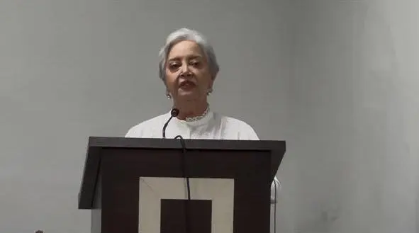 Mulher de cabelos brancos presos, brincos e blusa social branca discursa frontalmente atrás de um púlpito de madeira escura com um detalhe retangular claro na frente.