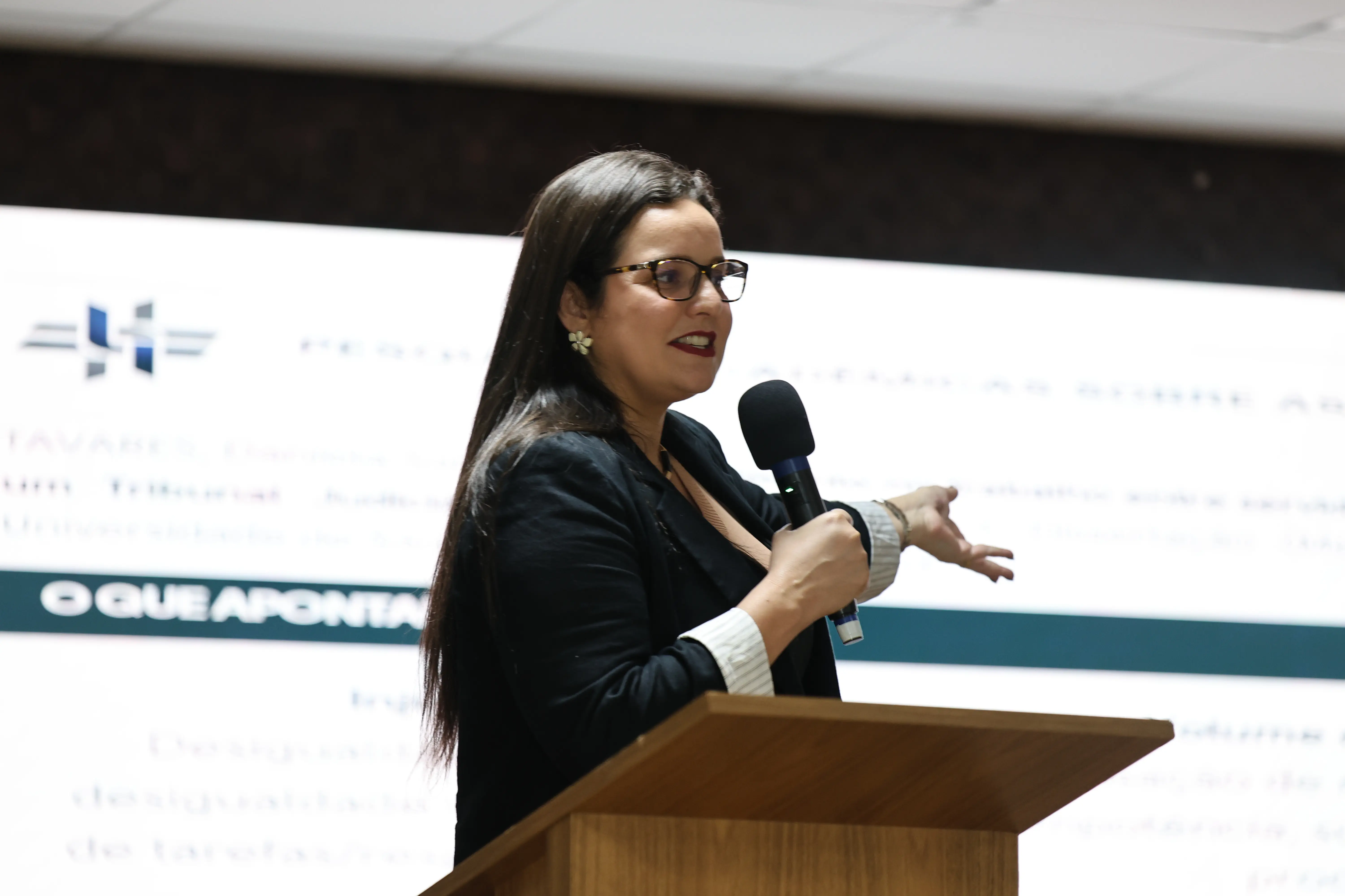 Uma mulher de cabelos escuros compridos, óculos e batom vermelho, veste blazer preto e fala ao microfone sobre um púlpito de madeira. Ao fundo, tela exibe slide da palestra.