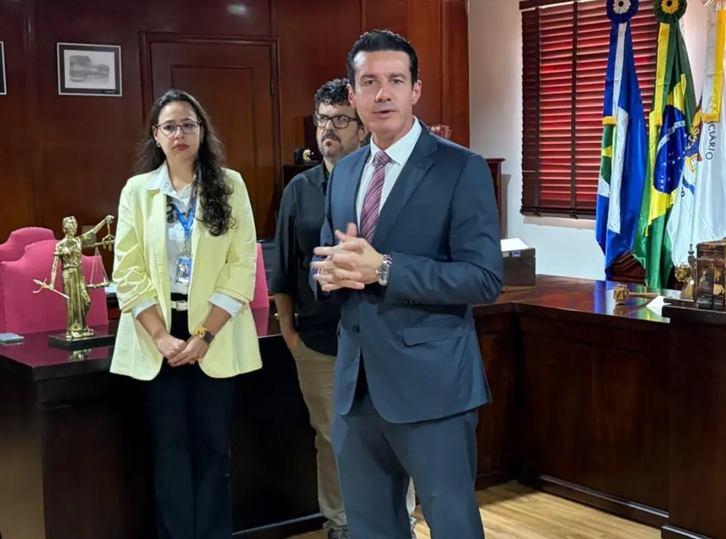 Homem de terno cinza e gravata listrada fala gesticulando com as mãos. Ao fundo, duas pessoas observam, e bandeiras do Brasil e de Mato Grosso compõem o cenário.