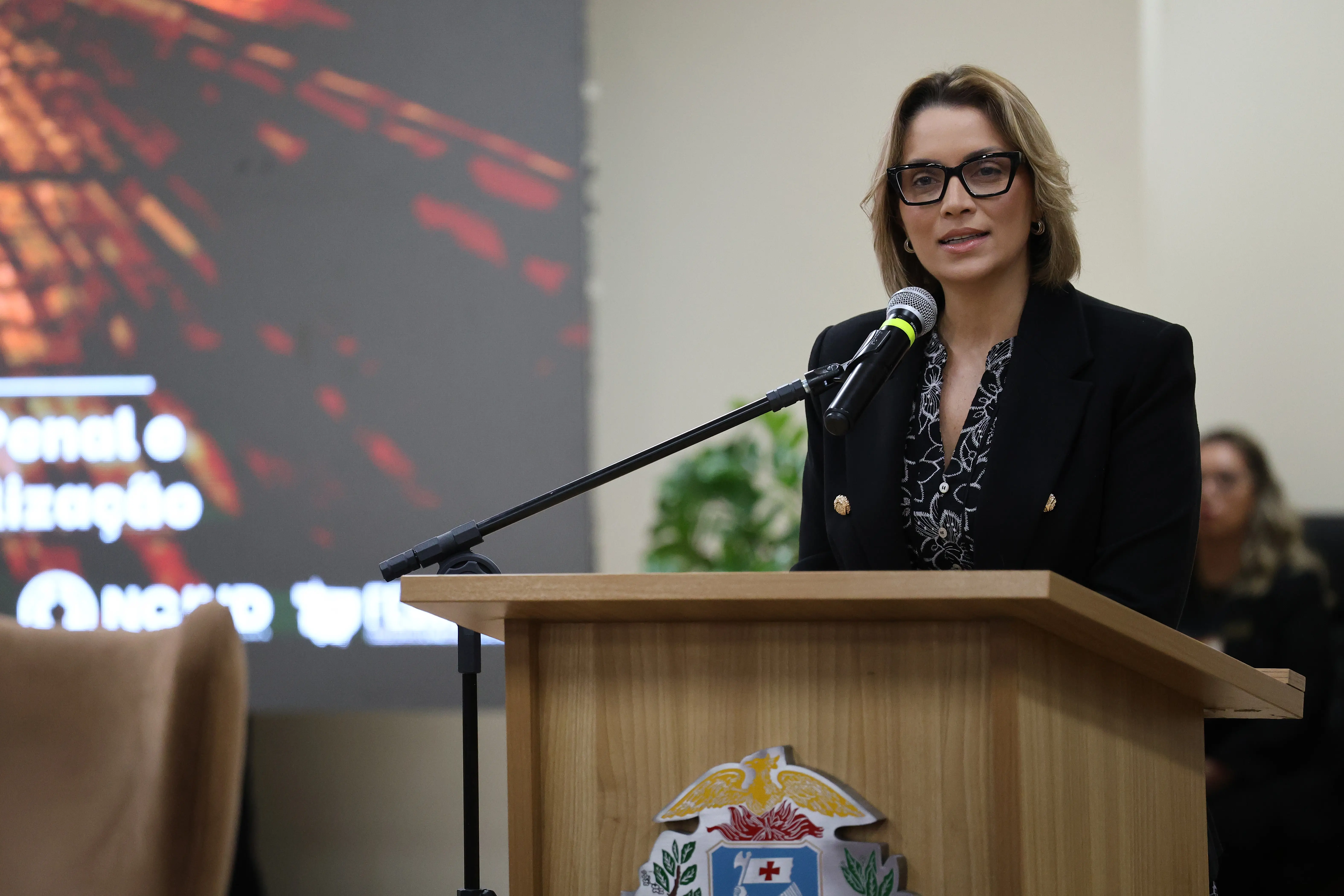 Mulher branca de óculos e blazer preto fala ao microfone em um púlpito com brasão do Mato Grosso. Ao fundo, há um telão com texto desfocado e uma pessoa sentada à direita.
