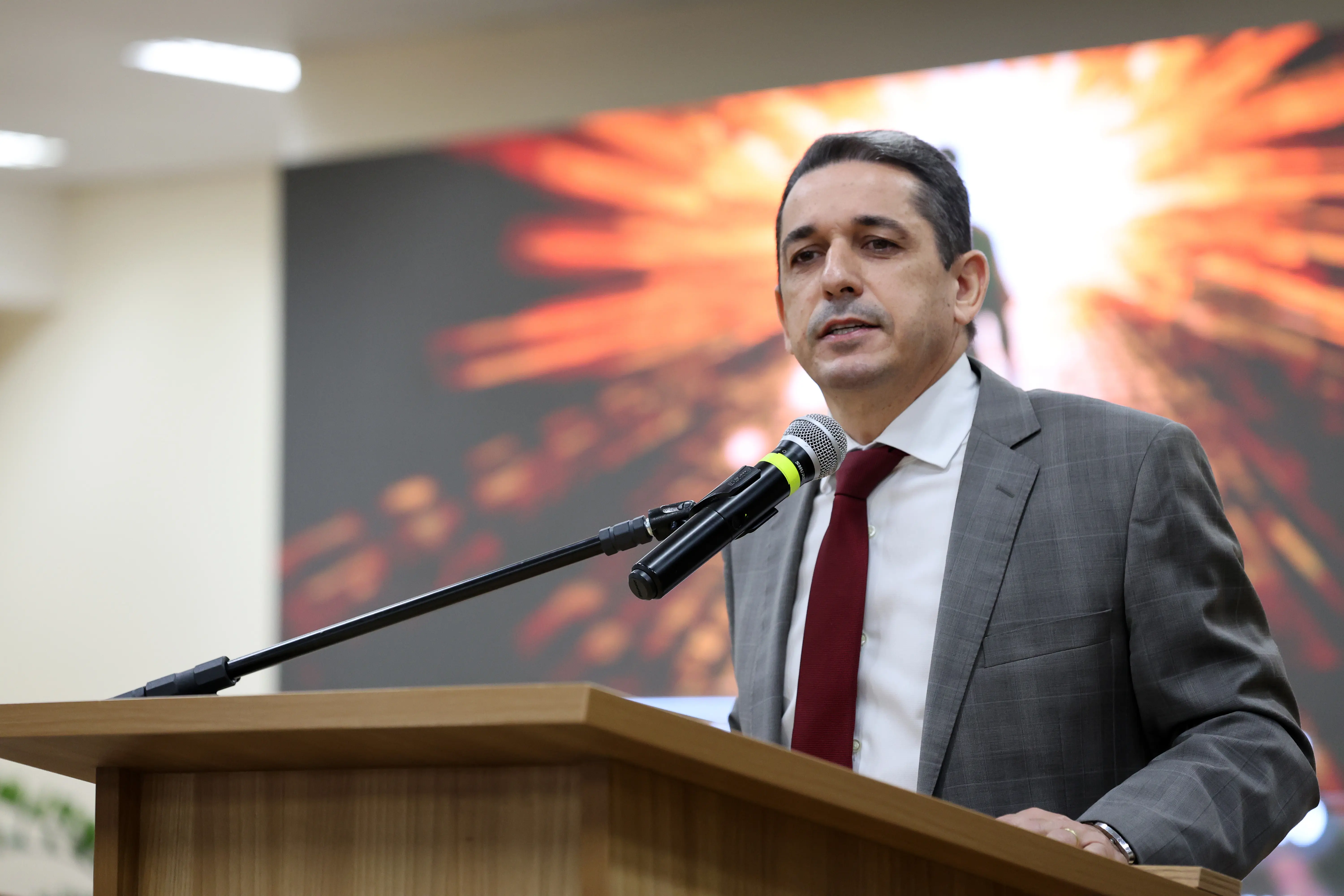 Homem branco de cabelos escuros fala ao microfone em um púlpito de madeira. Ele veste paletó cinza quadriculado, camisa branca e gravata vermelha. Ao fundo, um telão exibe uma imagem alaranjada.