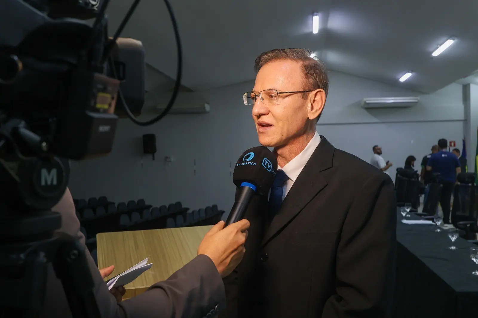 Homem branco de óculos e terno preto concede entrevista falando ao microfone da