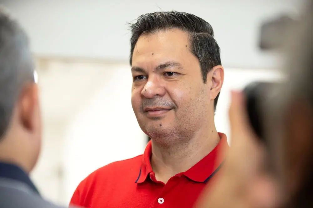Homem adulto de pele clara e cabelos escuros, vestindo uma camisa polo vermelha, sorri levemente enquanto conversa com pessoas ao seu redor. Ele está posicionado no centro da imagem, sendo visto de perfil parcial, com o foco voltado para o seu rosto. O plano de fundo é claro e levemente desfocado.