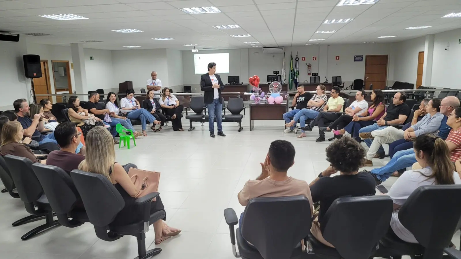 Foto mostra o juiz Ricardo Nicolino em pé, conduzindo o encontro e conversando com os participantes, que estão sentados em um círculo. Ao fundo, uma mesa decorada com balões em formato de coração e outros elementos alusivos ao tema da adoção compõe o ambiente.