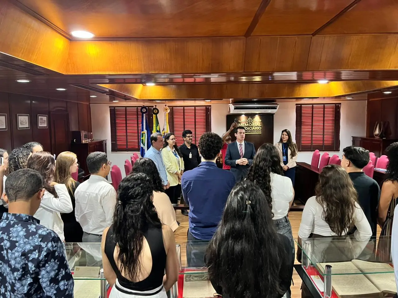 Grupo de jovens reúne-se ao redor de mesas com vitrines em sala do Memorial do Judiciário Mato-Grossense, enquanto homem de terno discursa ao centro.
