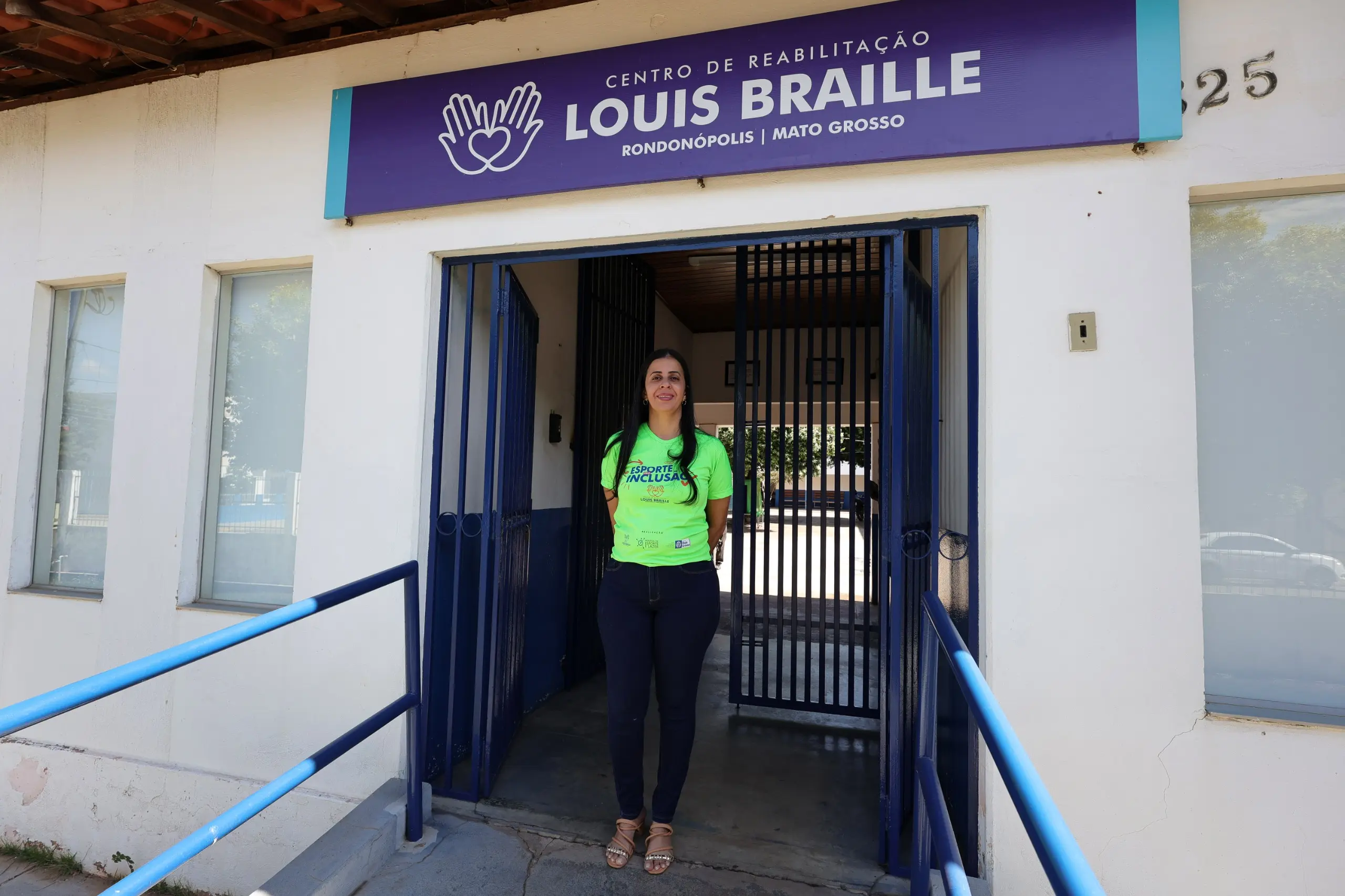 Mulher branca de cabelos pretos e longos, vestindo camiseta verde e calça escura, sorri em pé na entrada sob a placa Roxa escrito Centro de Reabilitação Louis Braille, Rondonópolis.