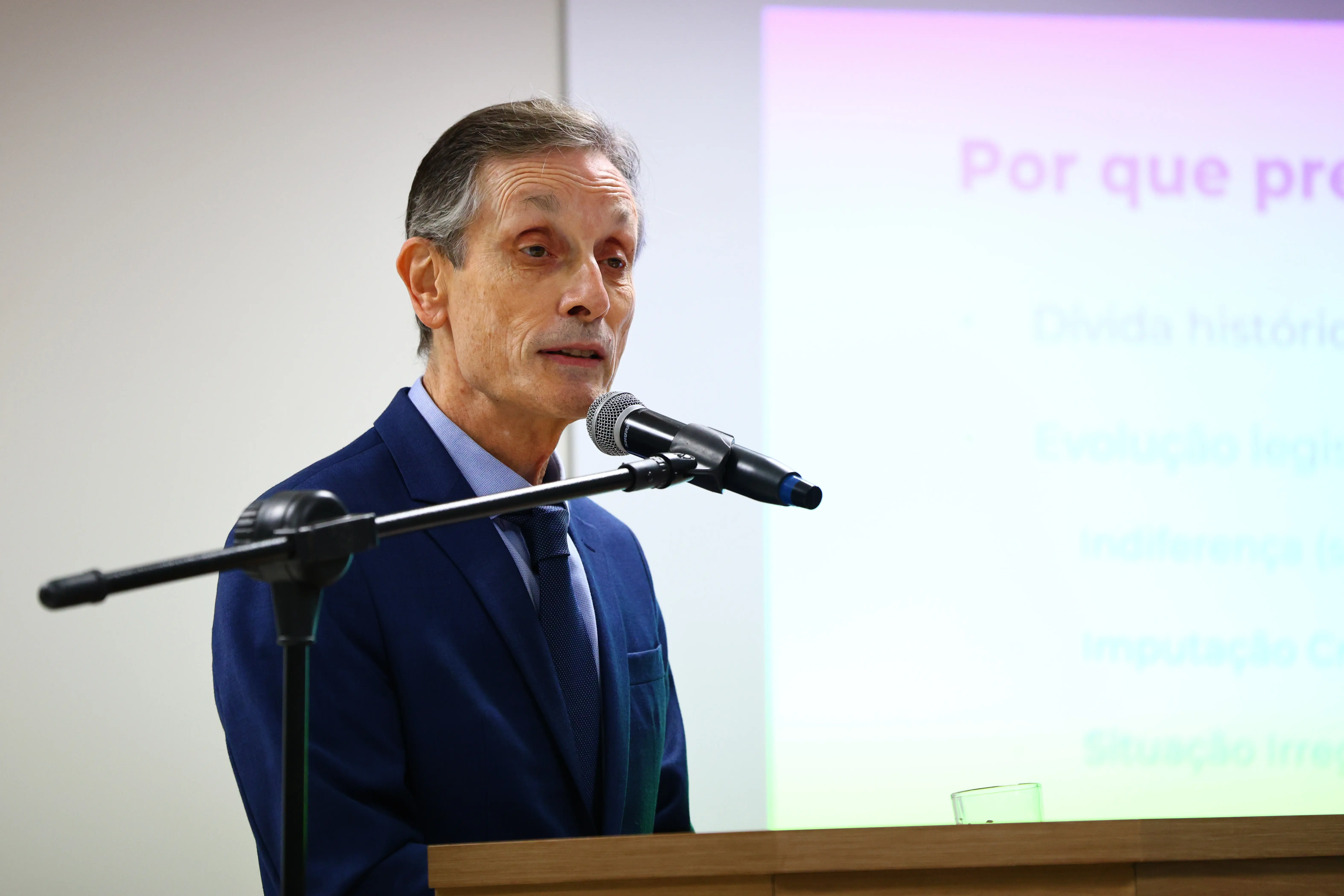 Homem de terno azul e gravata fala ao microfone em um púlpito de madeira. Ao fundo, uma tela de projeção clara exibe um texto desfocado com o título em rosa 