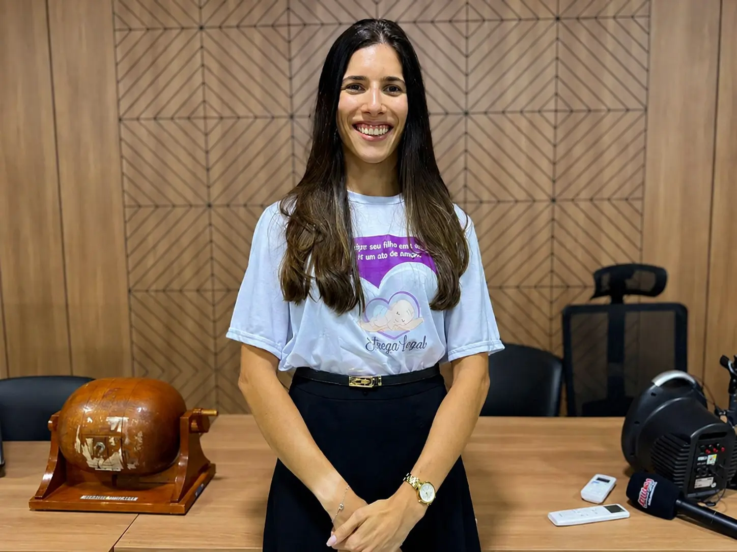 Foto posada da juíza Lais Paranhos. Ela está sorrindo em uma sala de reuniões, em frente a uma parede com revestimento geométrico amadeirado. Ela usa camiseta clara do programa Entrega Legal e saia preta. Sobre a mesa ao fundo há equipamentos de áudio e um objeto de madeira decorativo.