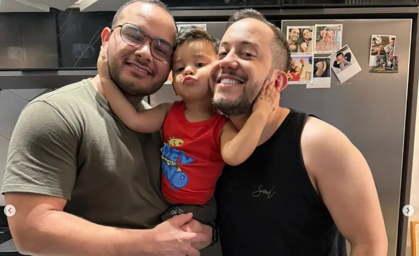 Dois homens sorridentes abraçam um menino de regata vermelha no centro, que faz bico para a foto. Eles estão em uma cozinha, posicionados em frente a uma geladeira cinza com fotos grudadas.