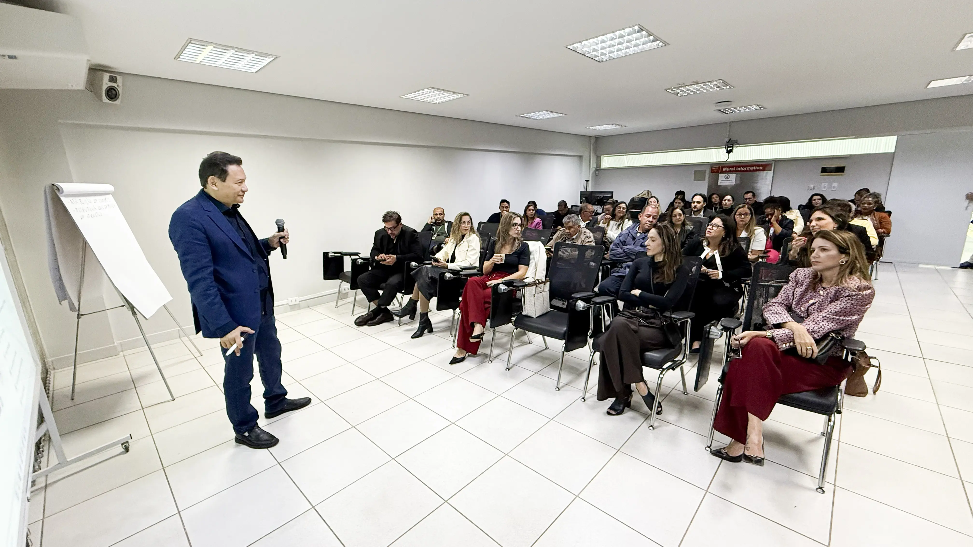 Dezenas de pessoas sentadas em cadeiras assistem a uma apresentação em sala institucional. Um homem de terno fala ao público, no microfone.