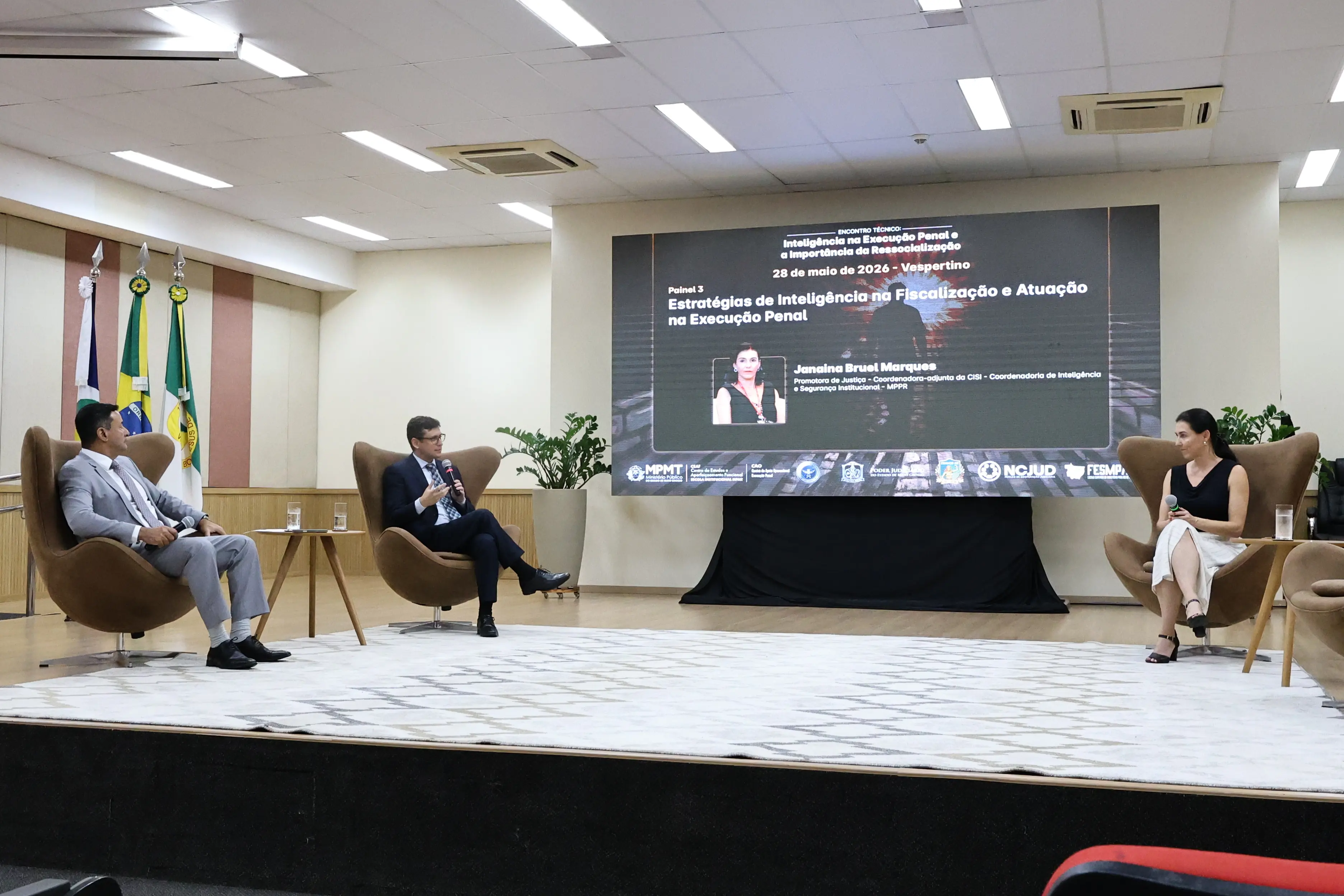 Três pessoas sentadas em poltronas bege sobre um palco debatem. Ao fundo, telão exibe o título 