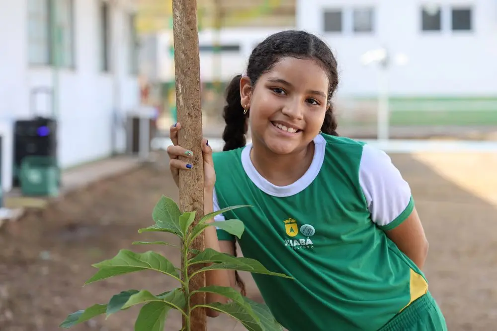 Foto horizontal que mostra a estudante Laís Bueno, de oito anos, sorrindo para a foto ao lado da muda de árvore plantada no pátio da escola municipal Dom Bosco. A menina usa uniforme verde e branco, ela tem a pele moreno, cabelos castanhos com duas tranças.