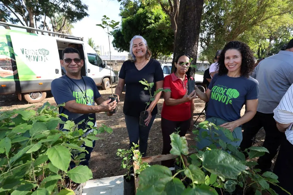 Foto horizontal em plano aberto que mostra um servidor e uma servidora com camiseta cinza do programa Verde Novo entregando mudas de planta para duas mulheres, no pátio da Escola Municipal Dom Bosco. Na frente deles, sobre uma mesa, há diversas mudas de plantas. Ao fundo, muitas árvores no pátio.