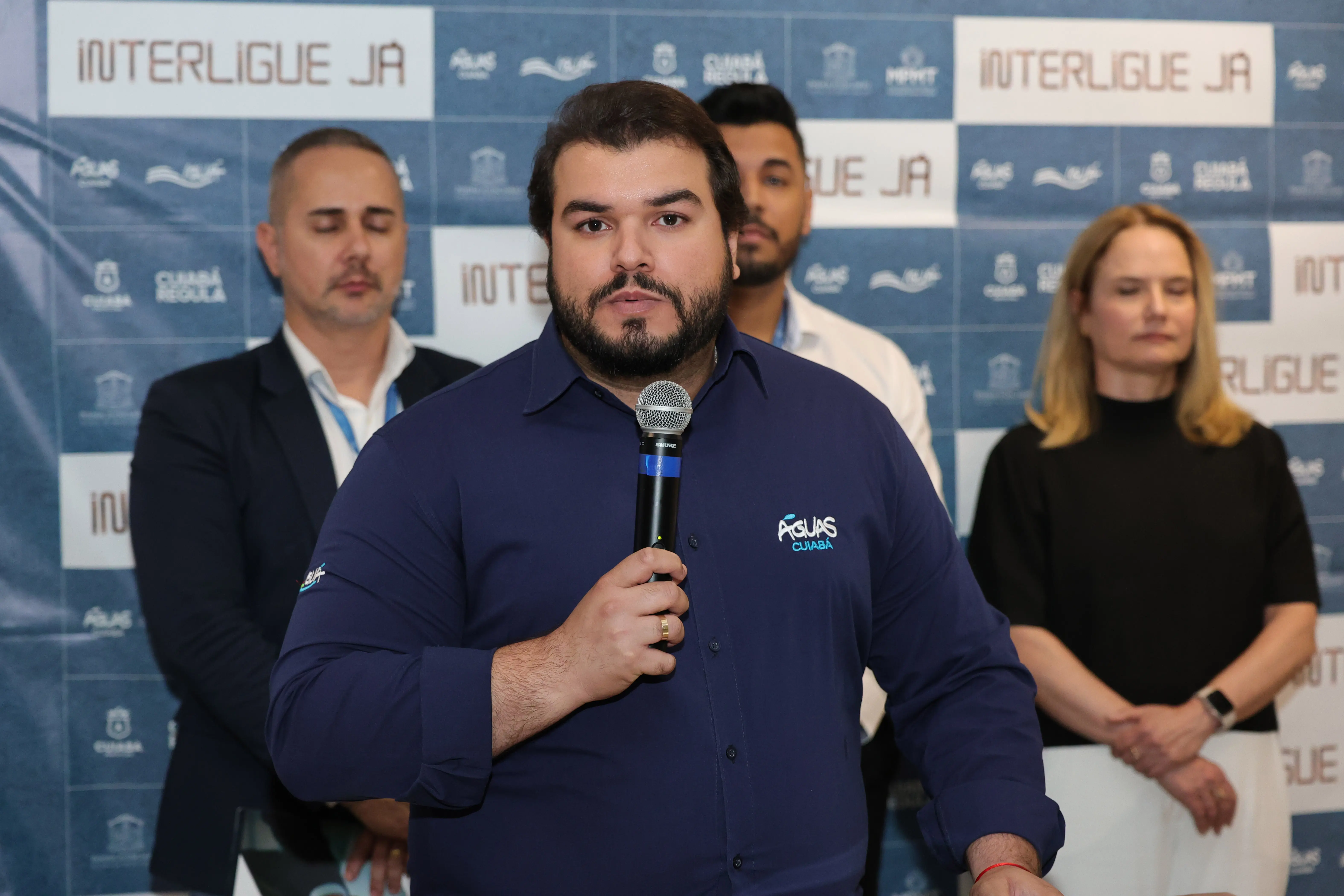 Homem de cabelos escuros e barba, veste camisa azul-marinho com logo 
