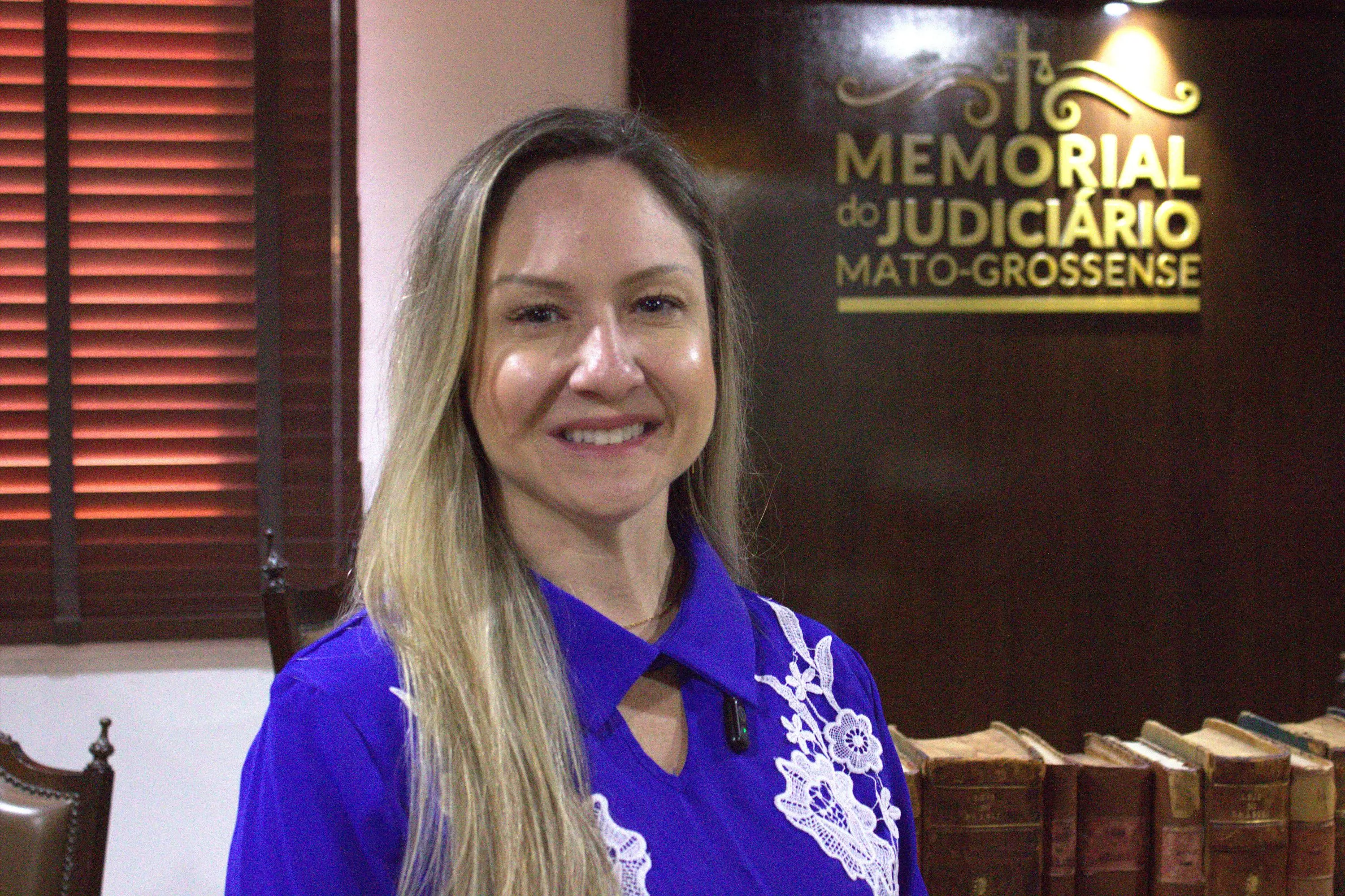 Mulher de cabelos longos loiros, blusa azul royal com renda branca, sorri para a câmera. Ao fundo, painel do Memorial do Judiciário Mato-Grossense.
