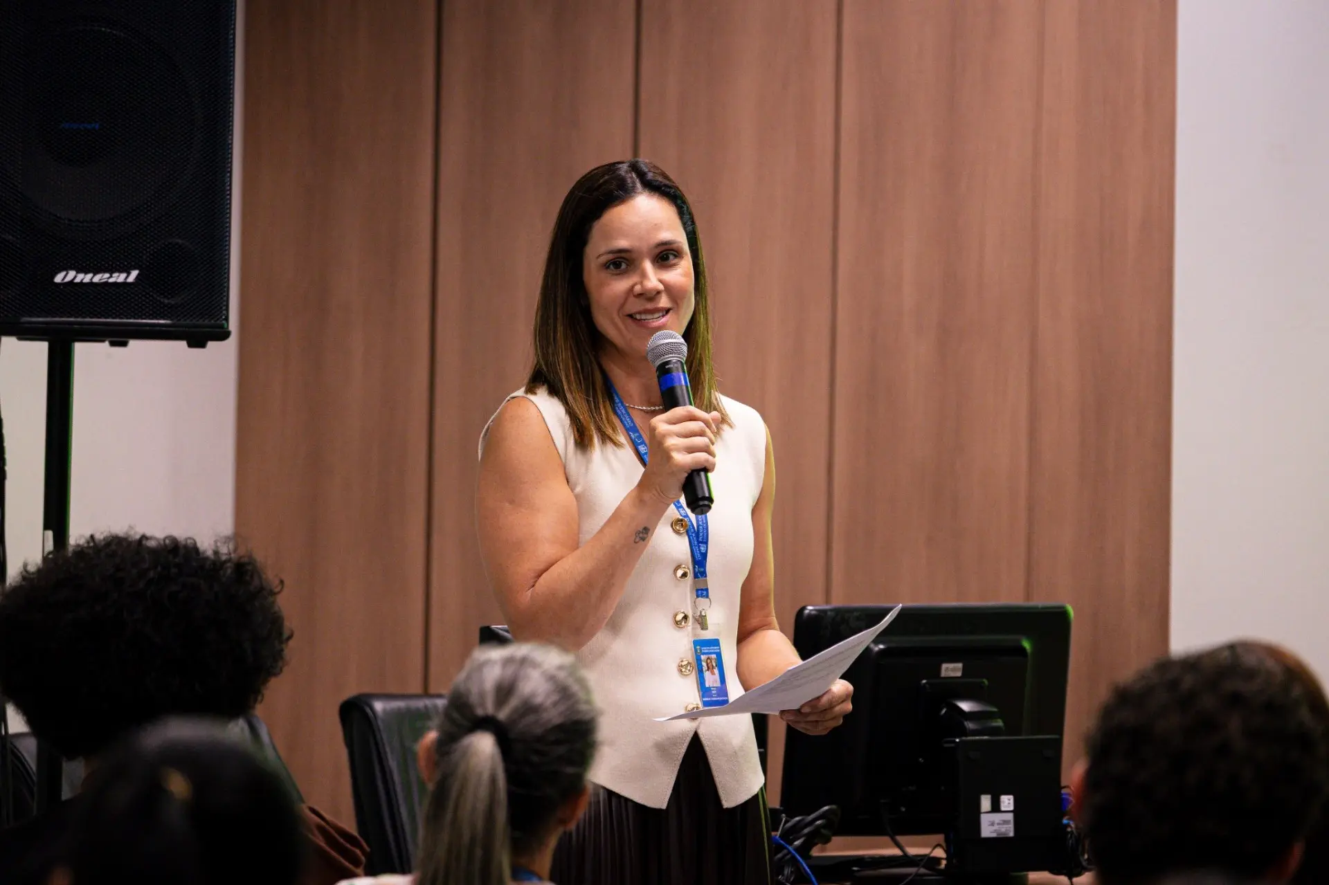 Foto horizontal que mostra a diretora do Daje, Shusiene Tassinari Machado, em pé, falando ao microfone para os estagiários que estão sentados. Ela é uma mulher branca, de cabelos castanhos claros, lisos na altura dos ombros, usando blusa estilo colete bege de botões na frente. 