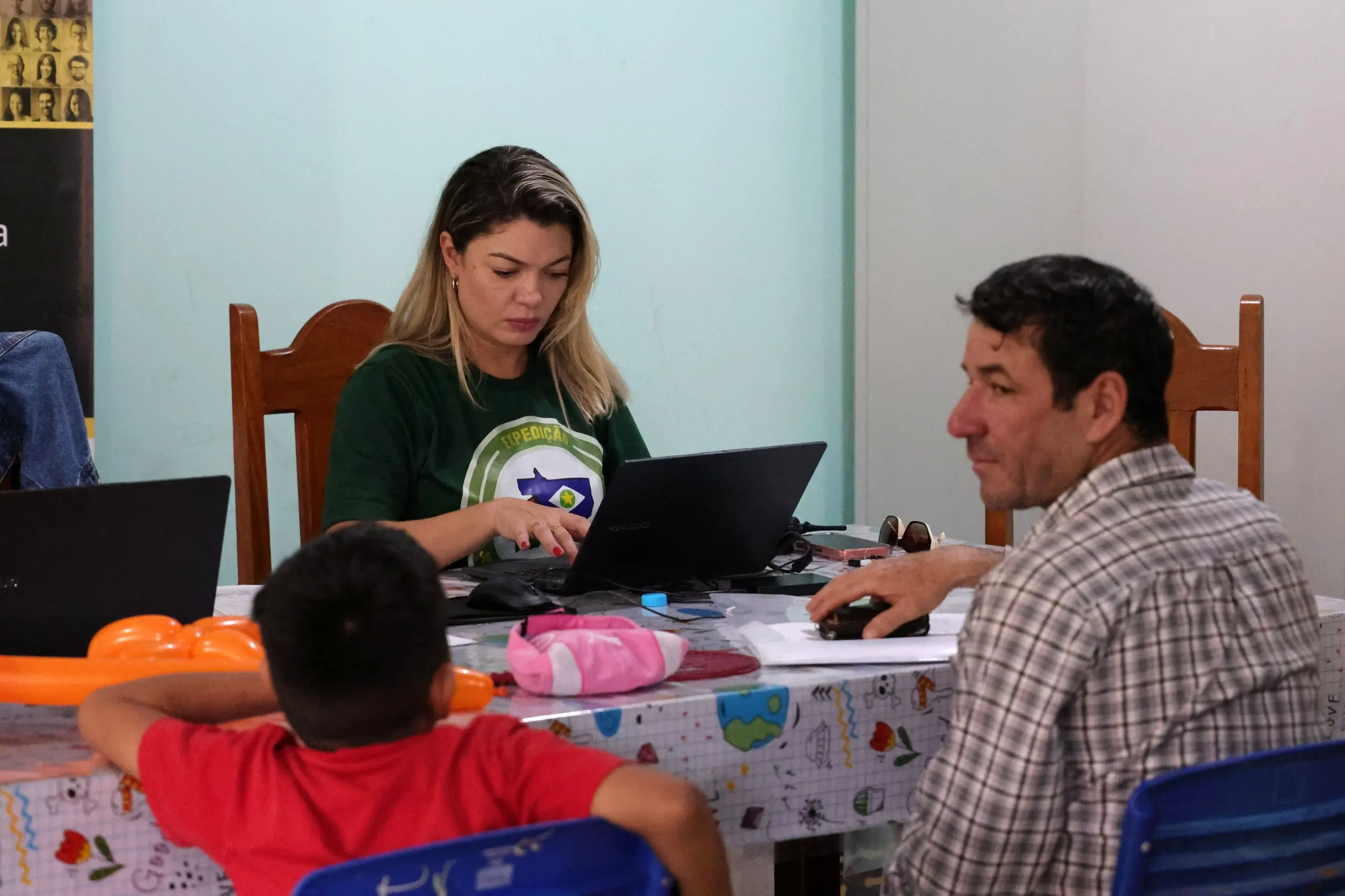 Mulher loira de camiseta verde digita no notebook à mesa. Diante dela, um homem de camisa xadrez de perfil e um menino de costas, com camiseta vermelha. Sobre a mesa, balões e objetos.