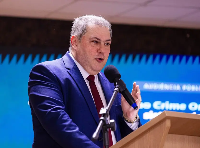 Homem de cabelos grisalhos curtos, terno azul e gravata vermelha fala ao microfone em um púlpito de madeira. Ao fundo, há um painel digital azul com a inscrição 