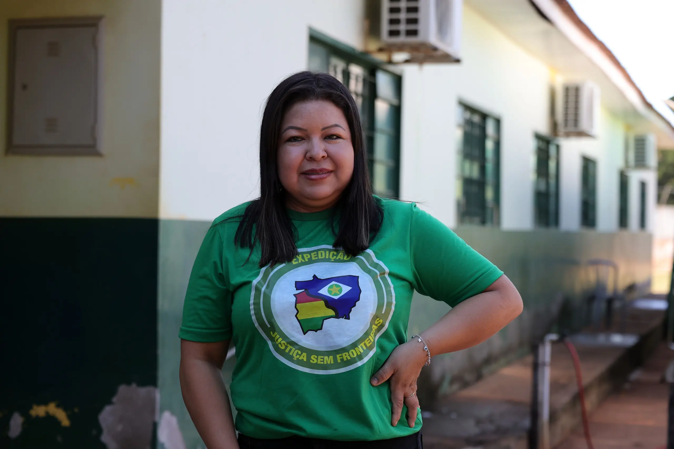 Mulher de cabelos escuros e lisos sorri para a foto, vestindo camiseta verde com o logotipo circular da 