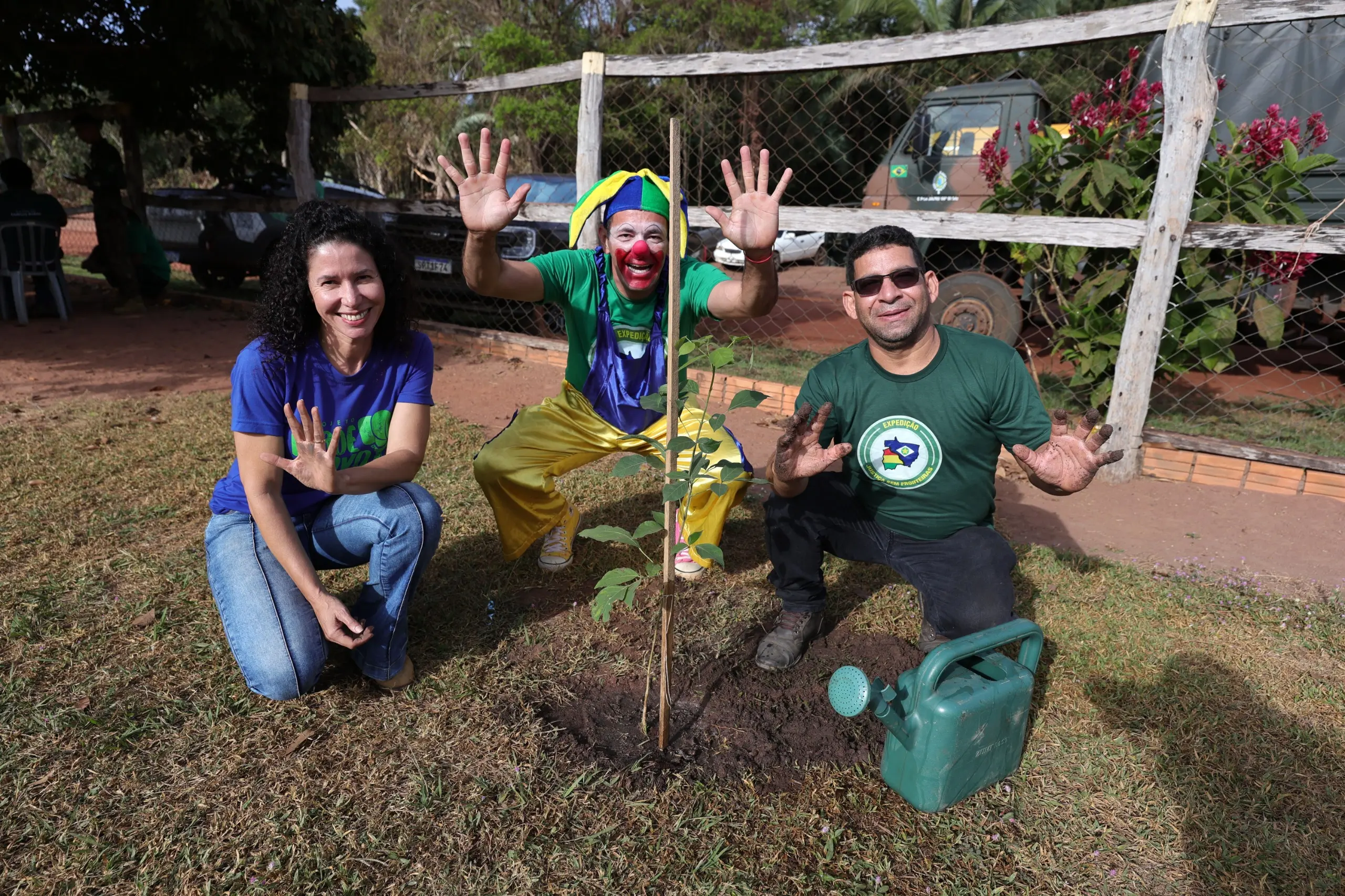 Três pessoas agachadas ao redor de uma muda de árvore: uma mulher de azul, um palhaço de verde e amarelo ao centro e um homem de verde à direita. Todos sorriem e mostram as mãos espalmadas.
