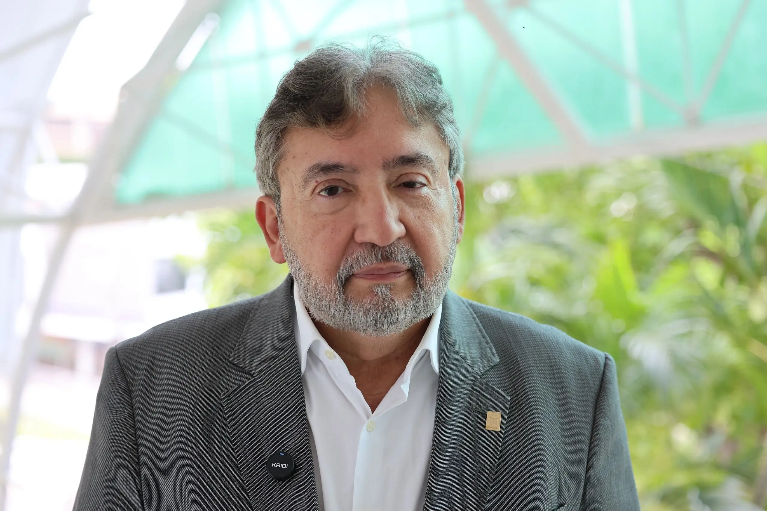 Homem de cabelos grisalhos e barba posa para a foto. Ele veste paletó cinza sobre camisa branca com um pequeno microfone de lapela preto preso à lapela. Ao fundo, plantas verdes e uma cobertura.