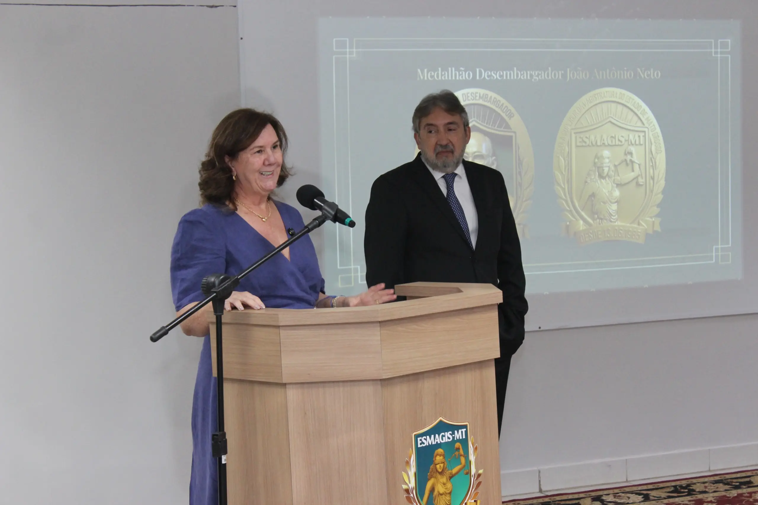 Mulher de vestido longo azul-escuro sorri e fala ao microfone em um púlpito de madeira clara com o brasão da ESMAGIS-MT. Ao lado direito dela, um homem de terno escuro, barba grisalha e gravata azul acompanha o discurso de pé. 