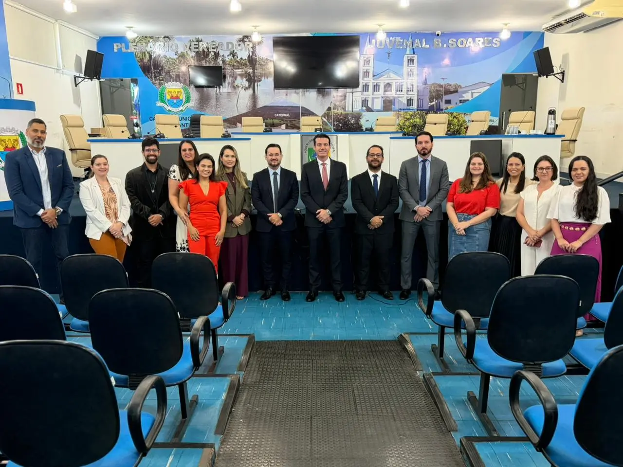 A imagem mostra o grupo de advogados da OAB que se reuniram com a equipe da Ouvidoria. O grupo tem quinze pessoas alinhadas em pé em frente ao palco do plenário da Câmara Municipal. Homens vestem ternos ou camisas sociais e mulheres usam vestidos, blusas e calças de cores variadas, como vermelho, br