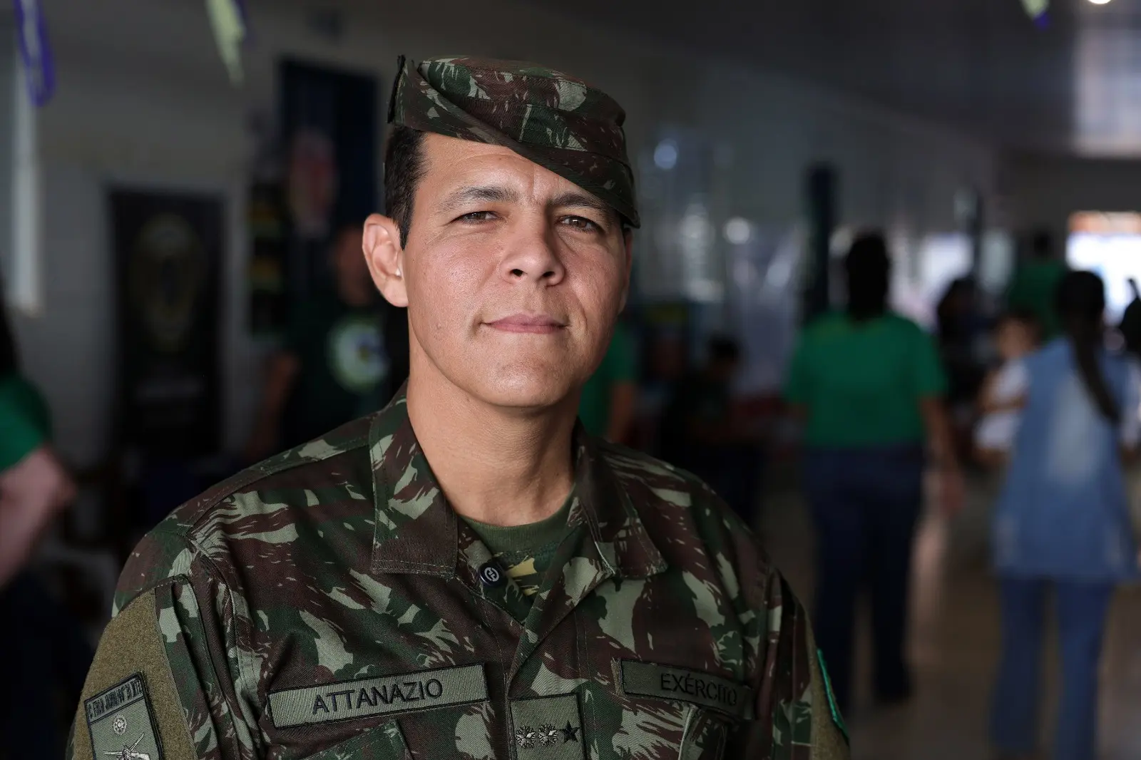 Homem de farda militar camuflada do Exército e boina olha sério para a câmera. Na farda há os nomes