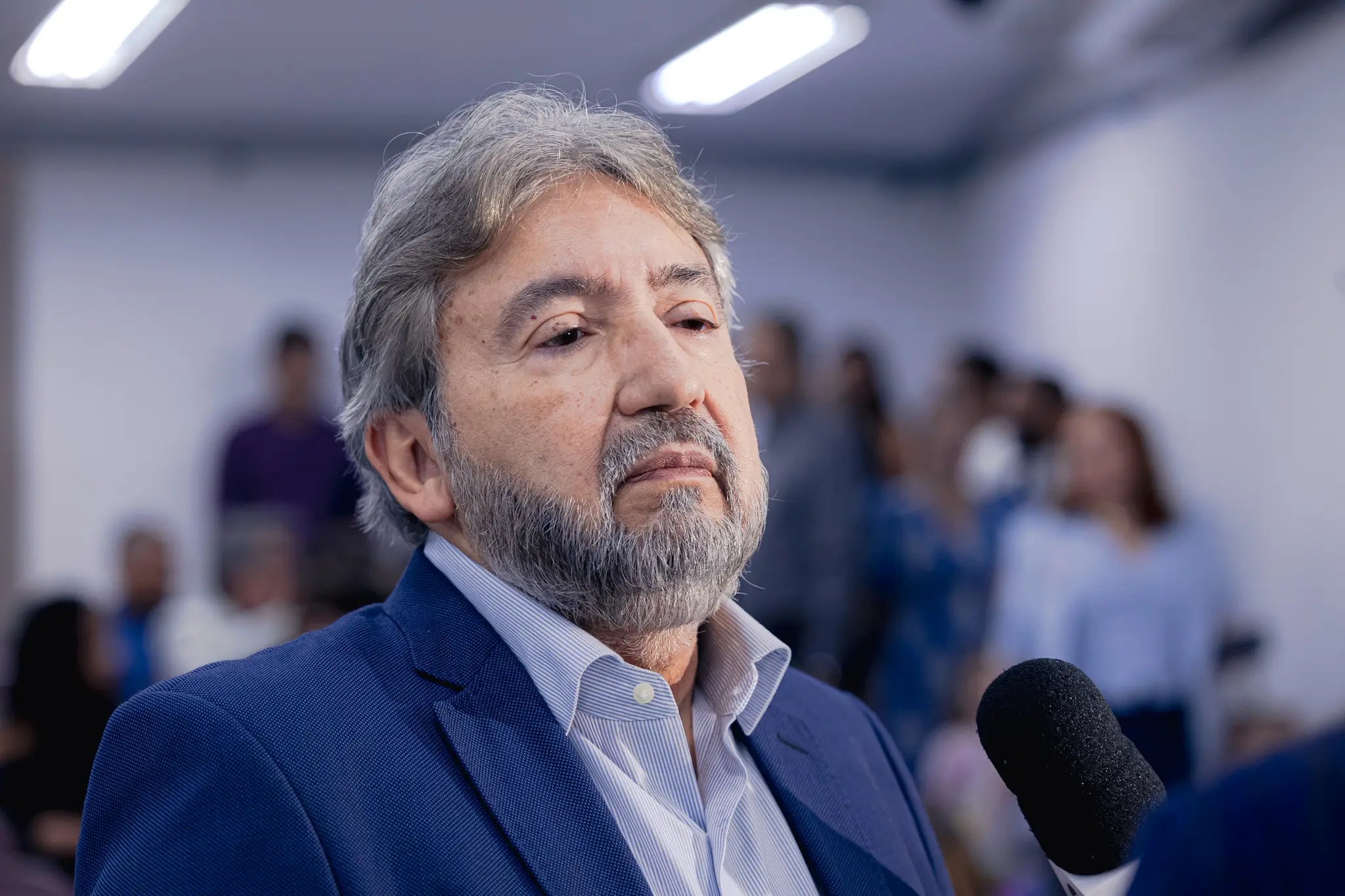 Homem grisalho e com barba, vestindo paletó azul e camisa listrada clara, concede entrevista com expressão séria. Um microfone preto aparece no canto inferior direito e o fundo está desfocado.