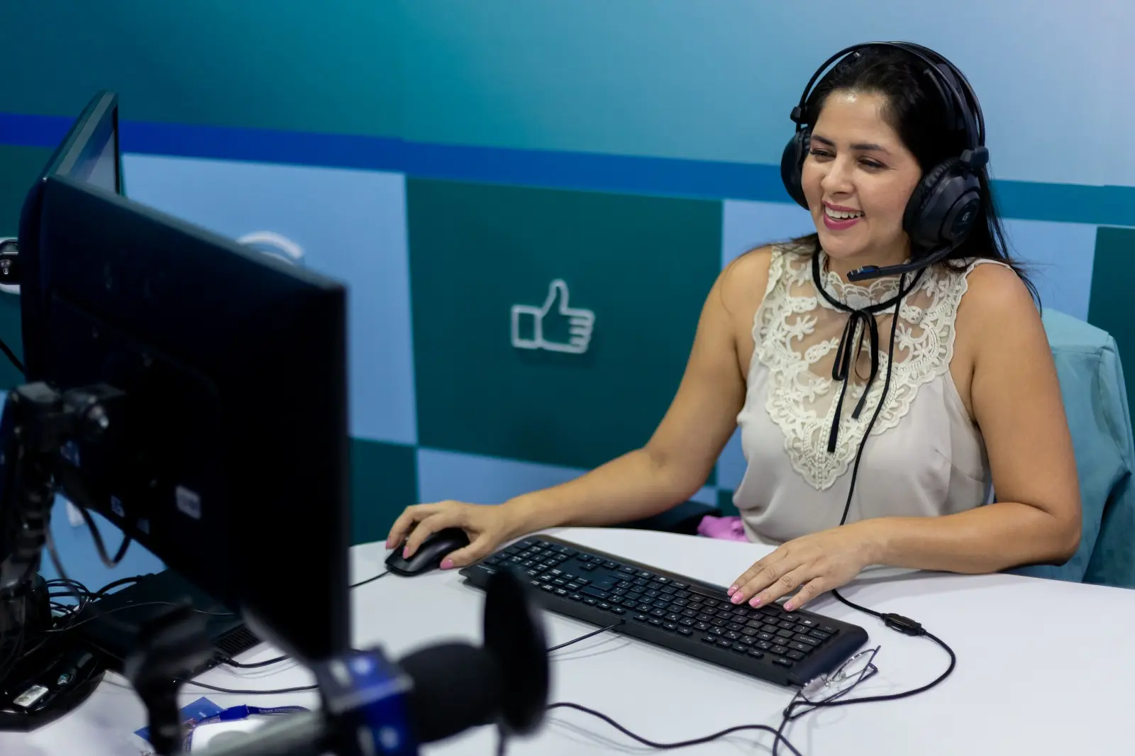 Mulher de blusa bege com detalhes em renda e um fone de ouvido headset sorri sentada em frente a um computador em um estúdio. Ela opera um mouse com a mão direita e um teclado com a esquerda. Ao fundo, uma parede verde e azul com o desenho de um
