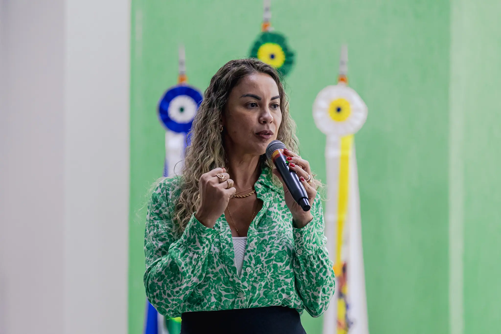 Mulher de cabelos longos e ondulados fala ao microfone, com a mão direita fechada. Veste camisa estampada verde e branca e saia escura. Ao fundo, parede verde e pontas de mastros de bandeiras.