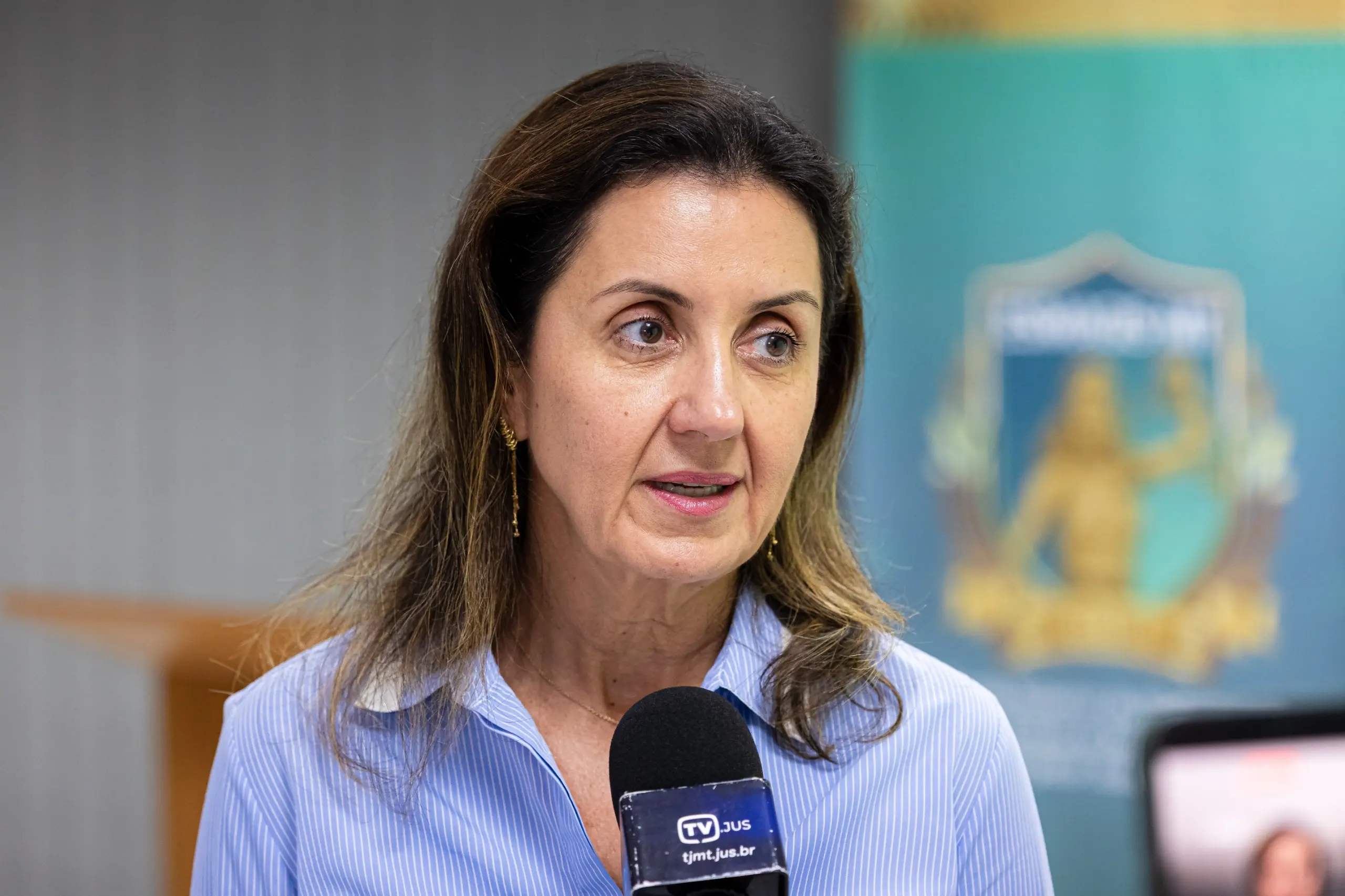 Uma mulher com cabelos castanhos na altura dos ombros e brincos dourados pendentes, está sendo entrevistada. Ela veste uma camisa social azul claro de botão e está sorrindo, olhando para alguém fora da câmera. Um microfone preto com um cubo de espuma azul claro e a inscrição 
