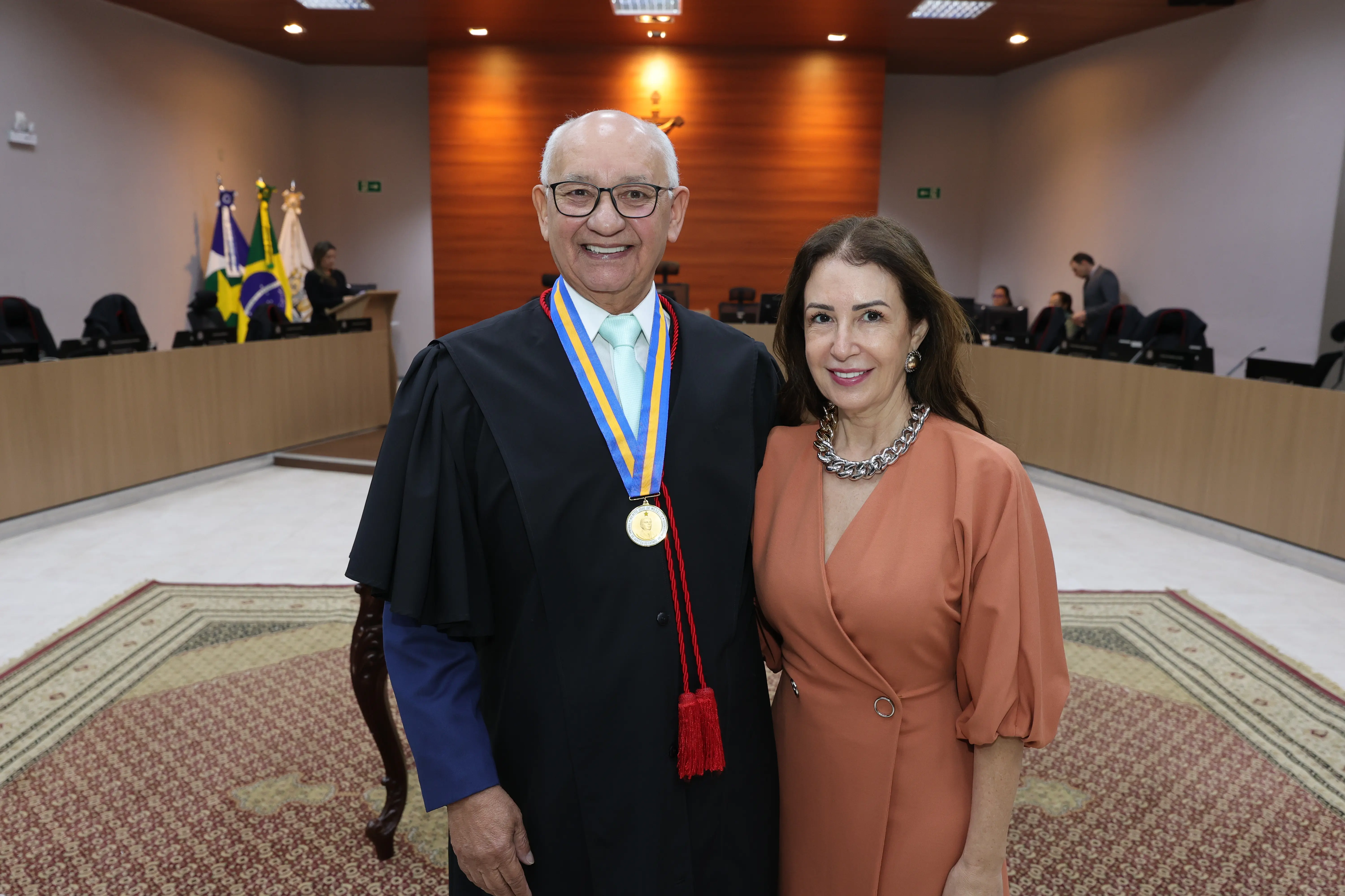 Desembargador Juvenal Pereira da Silva usa óculos e toga preta com medalha dourada sorri abraçado a sua companheira, Solange Wendt Ferreira, de cabelos longos e vestido longo coral. Eles estão em pé sobre um tapete estampado em um grande salão com mesas e pessoas ao fundo.