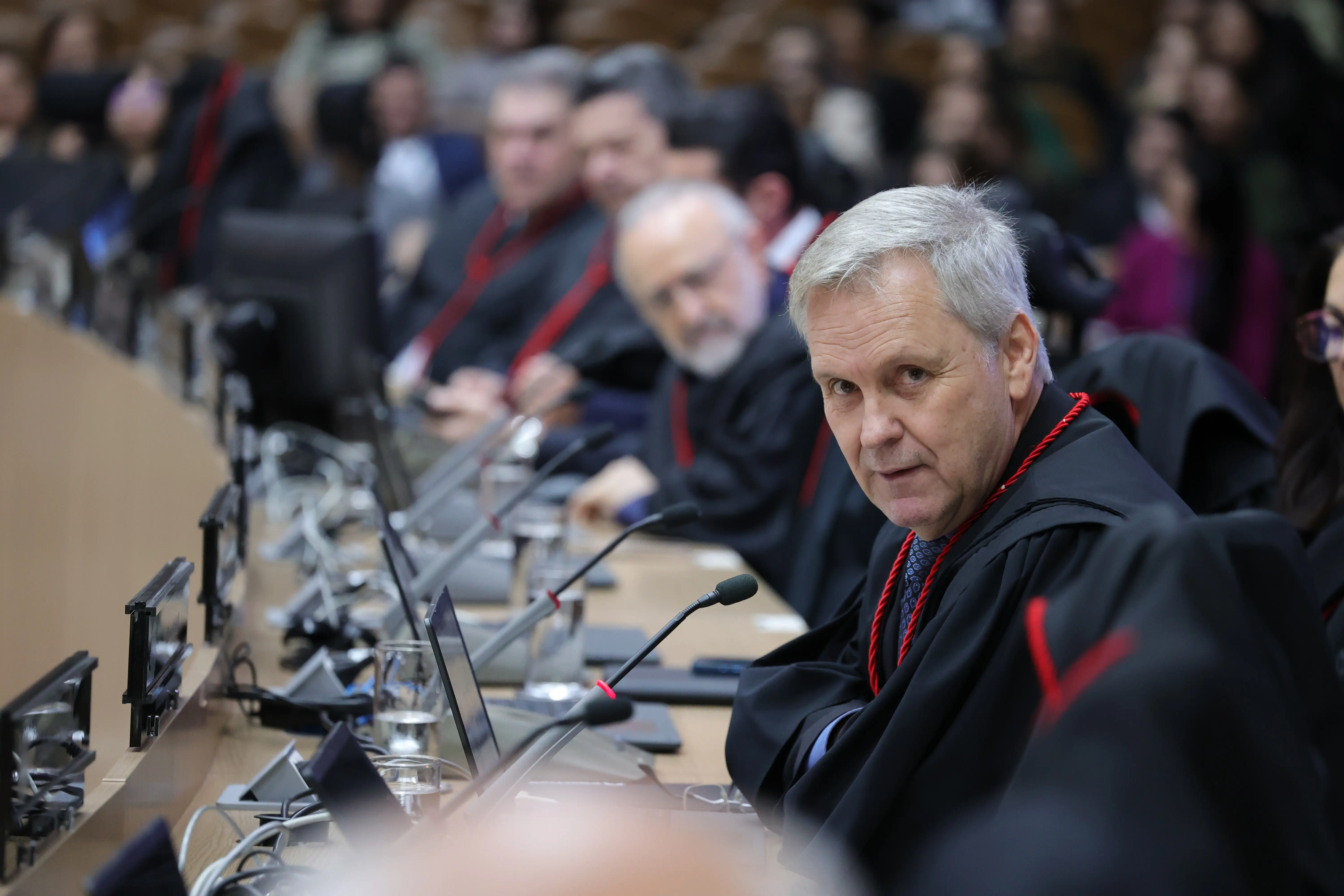 Fotografia do Desembargador Geraldo Giraldelli em primeiro plano, visto de perfil parcial, olha para a câmera. Ele veste uma toga preta com cordão vermelho e está sentado à mesa de um plenário. Ao fundo, uma fileira de outras autoridades vestidas de forma semelhante e uma plateia desfocada.