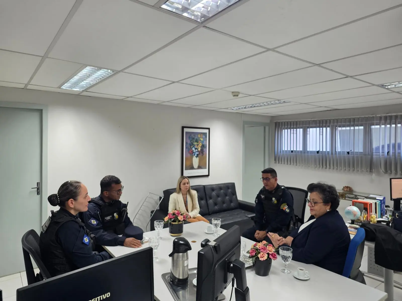 A imagem mostra cinco pessoas, entre as quais dois homens e uma mulher vestidos com uniforme azul da PMMT, e duas mulheres vestindo roupas formais. Todos estão sentados em torno de uma mesa, num gabinete administrativo, com computadores, flores e materiais de escritório.