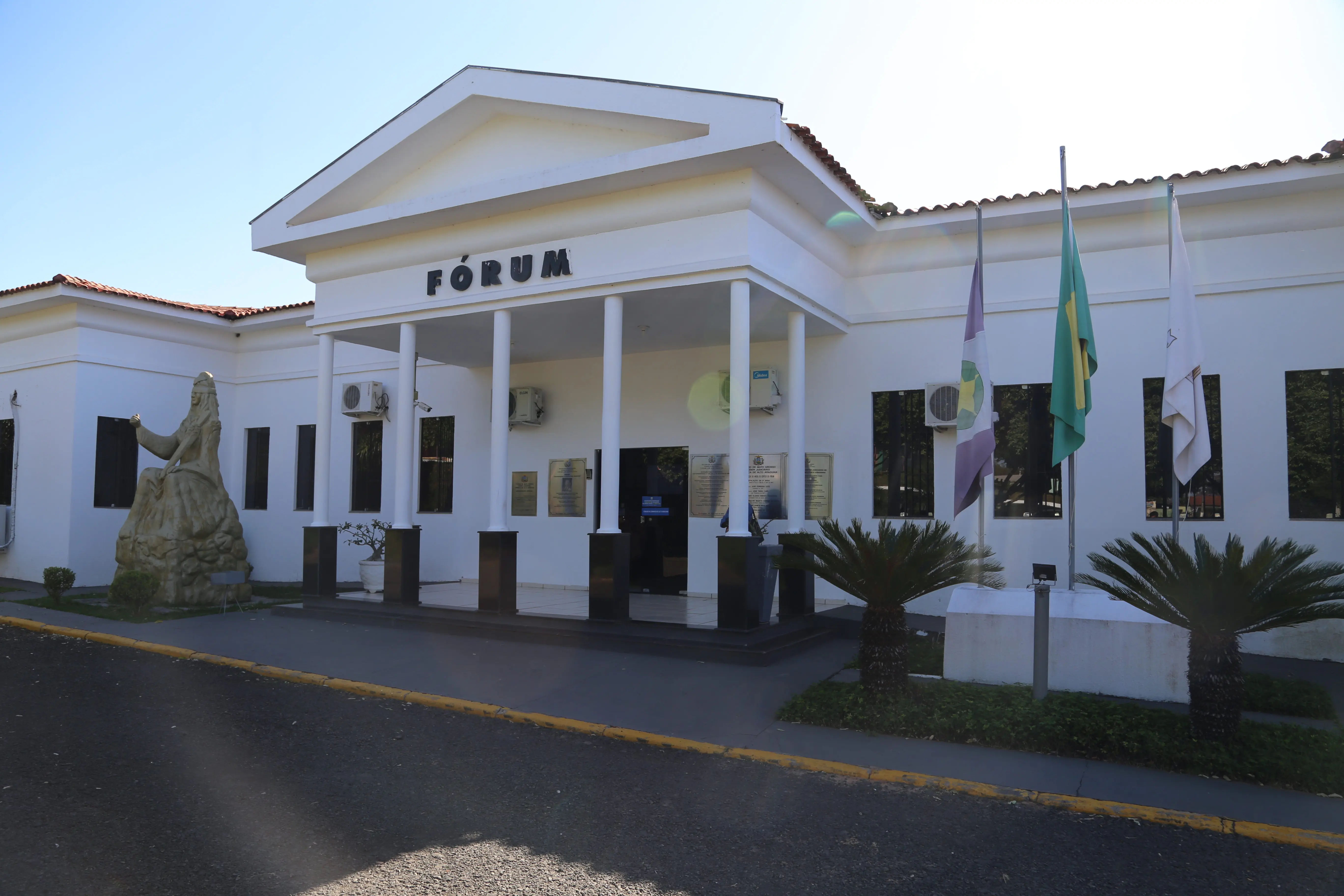 Fórum de Alto Araguaia, localizado em um prédio branco, com letras pretas, com bandeiras posicionadas à frente do espaço. 