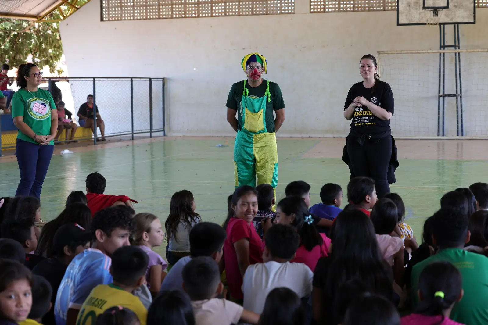 Na imagem um palhaço de macacão verde e amarelo e uma mulher de preto falam ao microfone para um grupo de crianças sentadas no chão de uma quadra poliesportiva coberta.