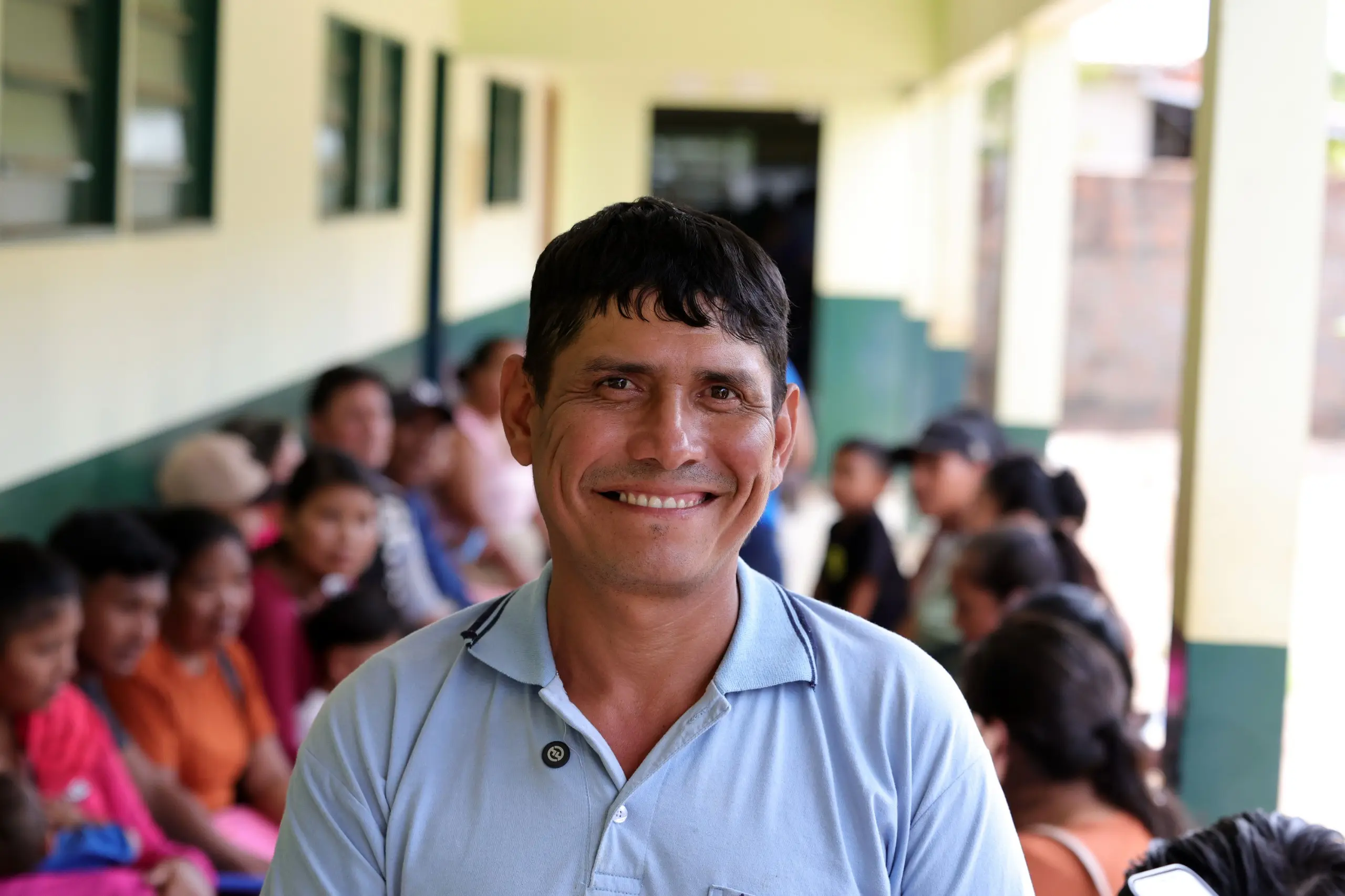 Foto horizontal que mostra o boliviano Gustavo Soliz sorrindo para a foto, em pé no corredor de uma escola, onde ocorreu o mutirão Justiça Sem Fronteiras. Ele é um homem com traços bolivianos, pele marrom, cabelos pretos e lisos, usando camisa polo azul celeste. 