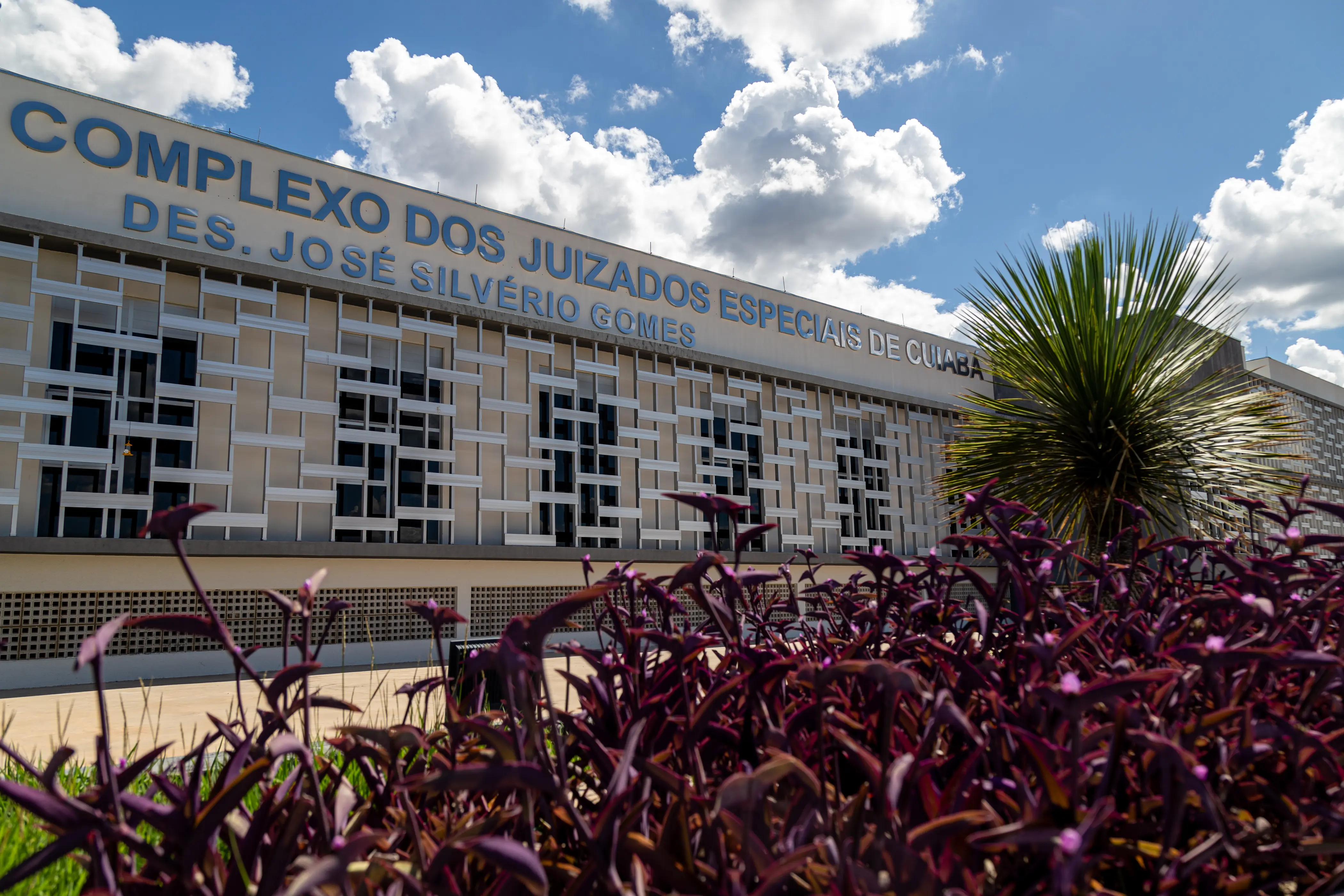 Fachada moderna do Complexo dos Juizados Especiais Desembargador José Ferreira Leite, em Cuiabá. O prédio tem estrutura de vidro e metal, com letreiro em destaque e jardim colorido com plantas roxas e verdes ao redor.