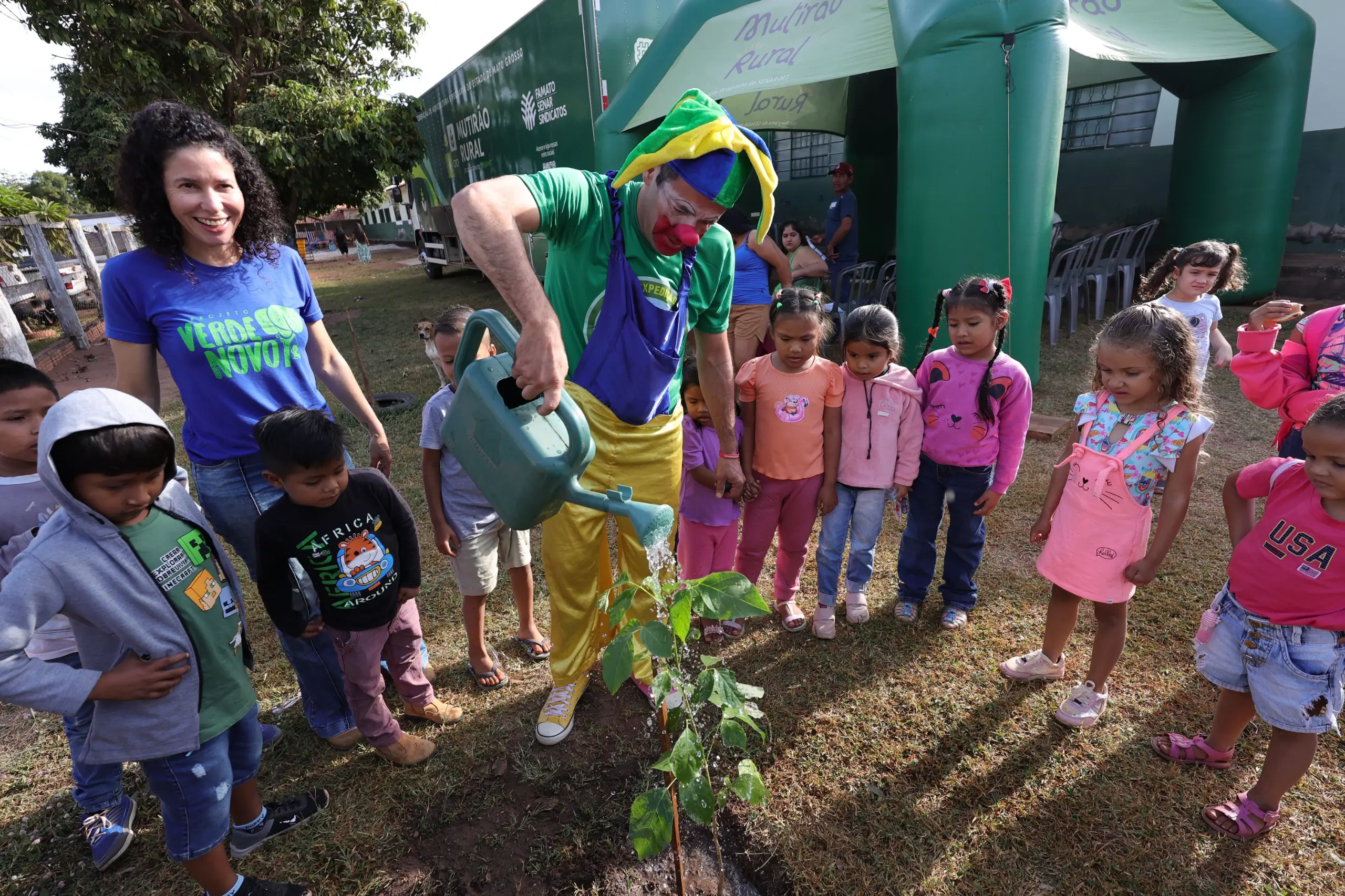 Palhaço com roupa verde e amarela rega uma muda de árvore com um regador verde. Uma mulher sorridente e várias crianças ao redor observam a ação em um espaço externo gramado.