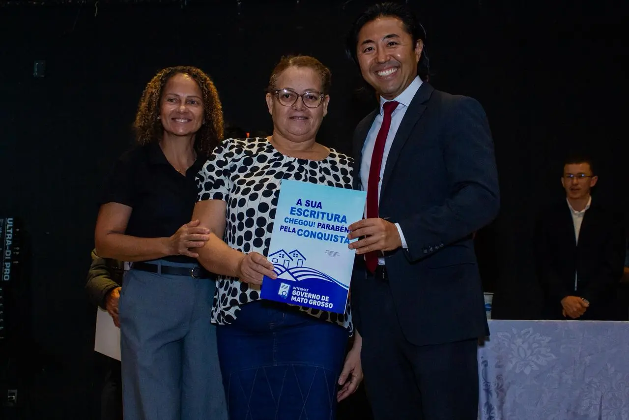 O juiz diretor do Fórum da Comarca de Jaciara, Fernando Kendi Ishikawa, posa ao lado de uma moradora durante a entrega de título definitivo de propriedade. A beneficiária segura o documento da propriedade nas mãos.