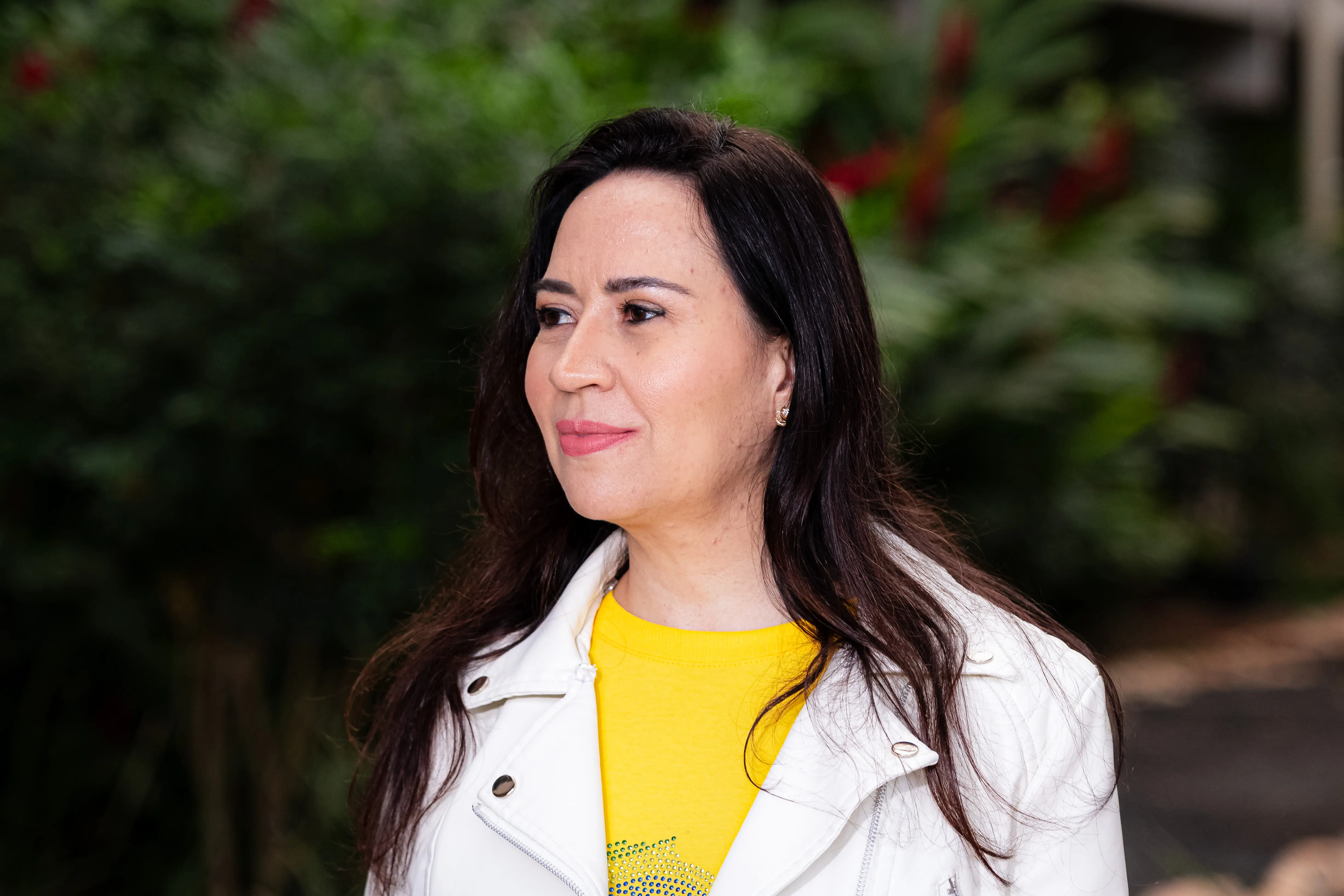 Mulher de cabelos castanhos ondulados, veste jaqueta branca sobre camiseta amarela com estrela, olha para o lado. Ao fundo, árvores com flores vermelhas.