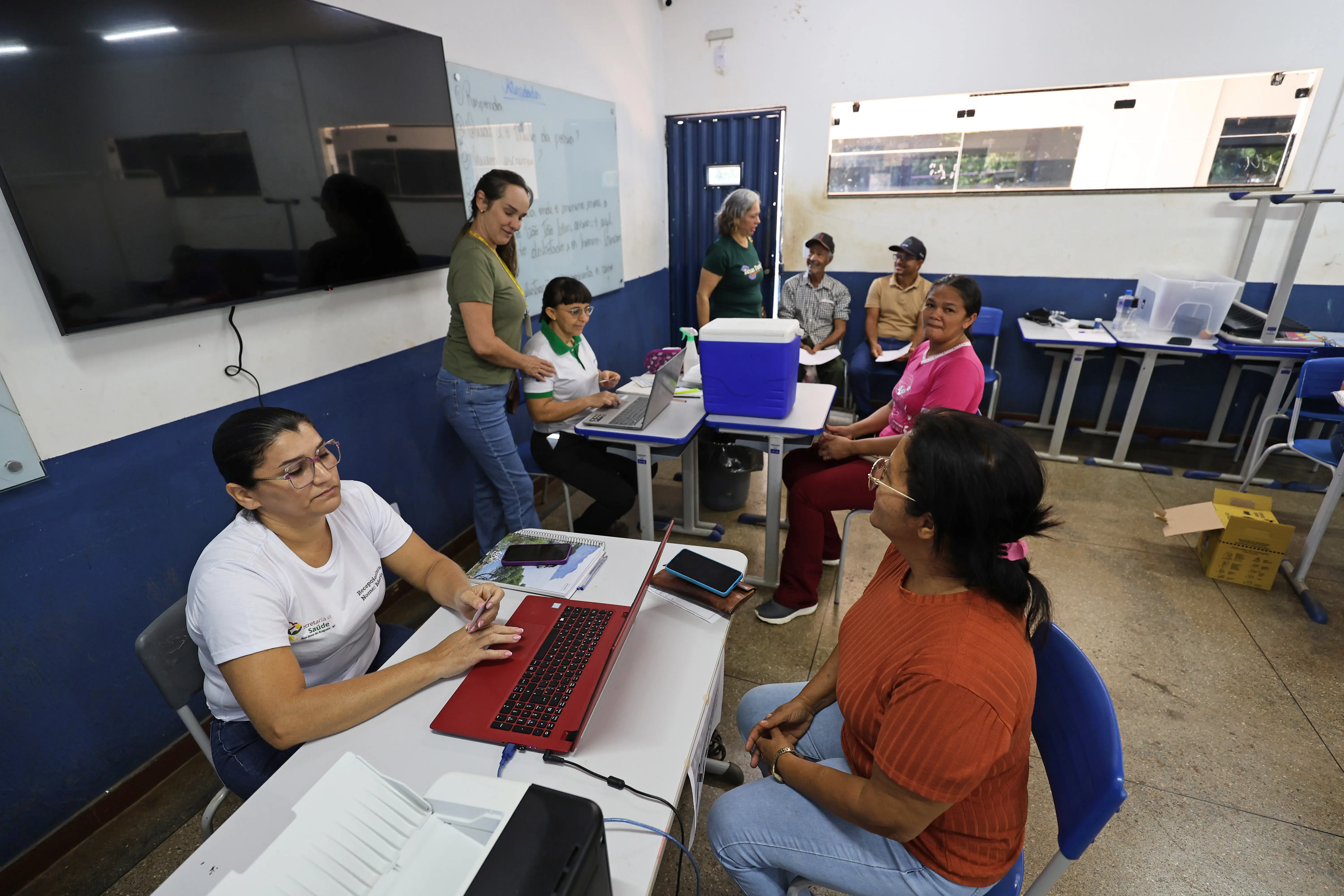 Sala de atendimento da Expedição Araguaia-Xingu reúne profissionais e moradores em ambiente simples, com paredes azul e branca e carteiras escolares. Mulheres atendem com notebooks, enquanto pacientes aguardam sorridentes.