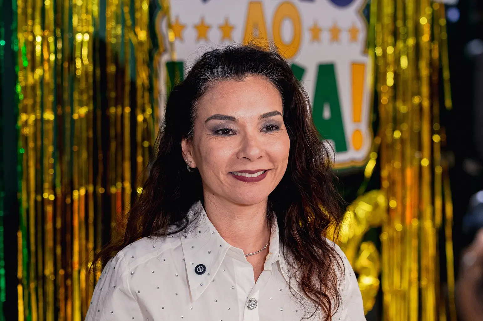 A juíza Hanae Yamamura de Oliveira posa para fotografia diante de uma decoração temática nas cores verde e amarelo. A magistrada veste camisa branca com detalhes discretos brilhantes e sorri para a câmera, tendo ao fundo elementos decorativos relacionados à Copa do Judiciário