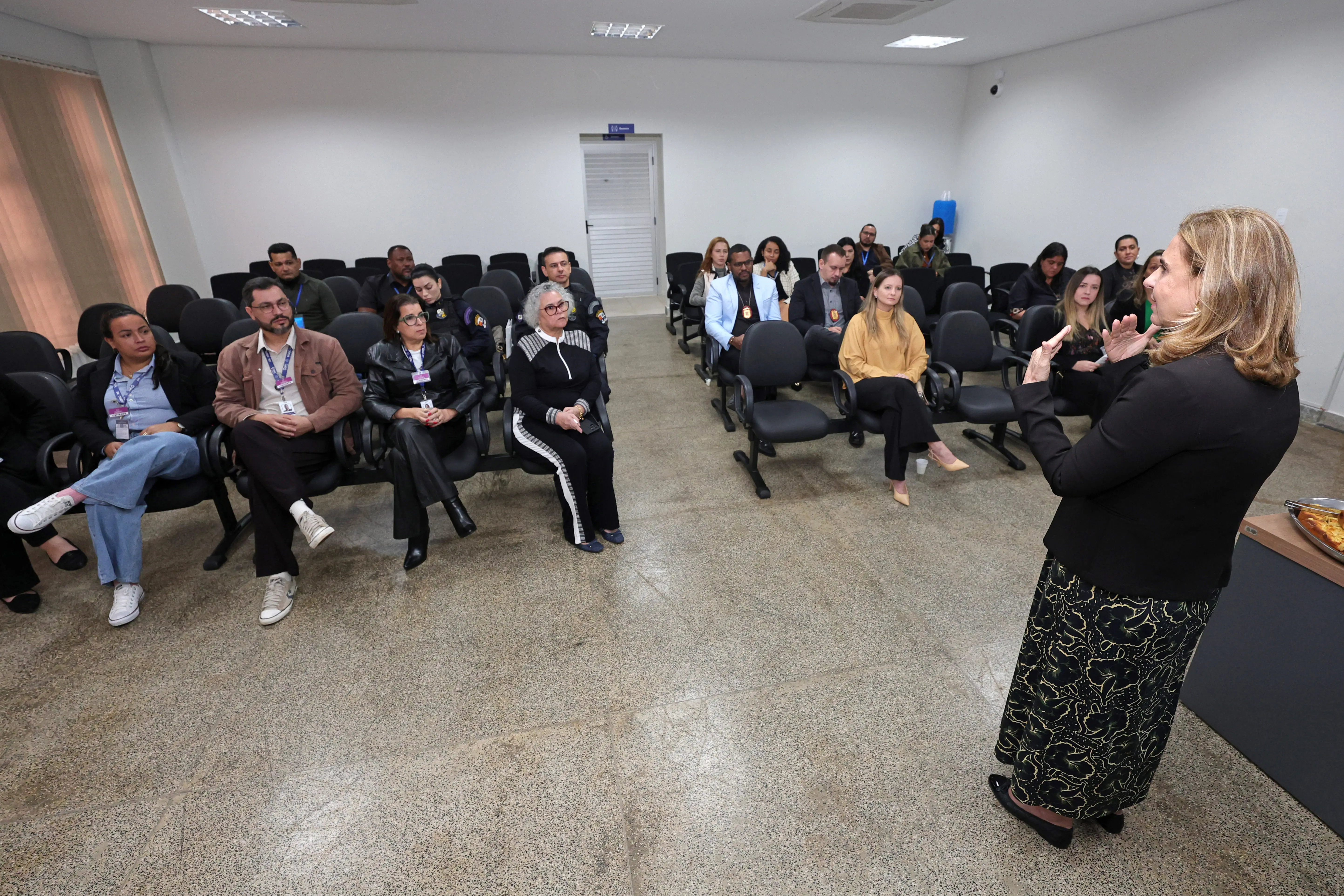 Mulher em pé, de costas para a câmera, fala e gesticula para uma plateia de homens e mulheres sentados em cadeiras escuras dispostas em fileiras, dentro de um auditório com paredes brancas