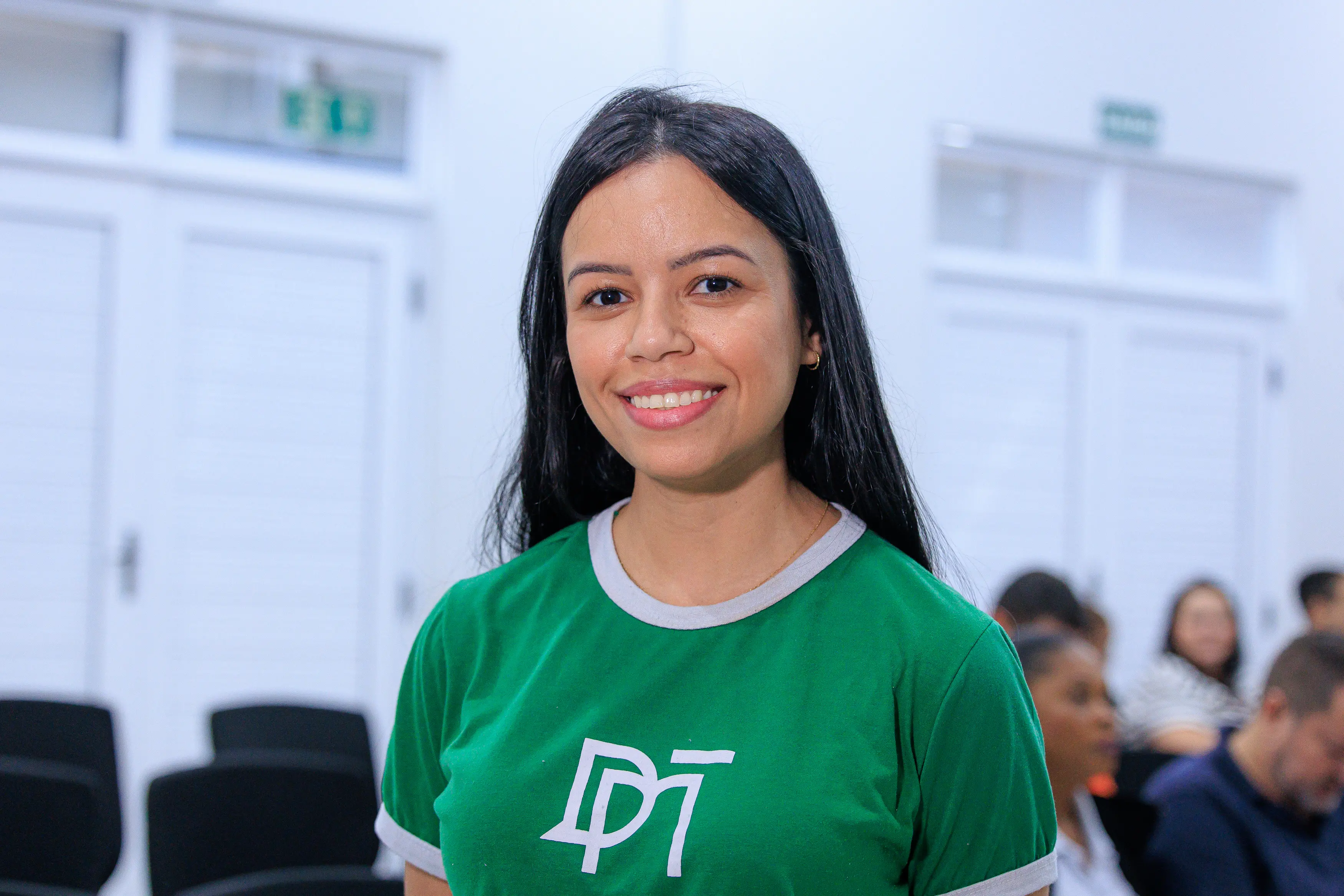 Servidora da Defensoria Pública, Wélida Lima é uma jovem com longos cabelos pretos sorri diretamente para a câmera. Ela veste uma camiseta verde-bandeira com a gola e as mangas brancas e o logotipo